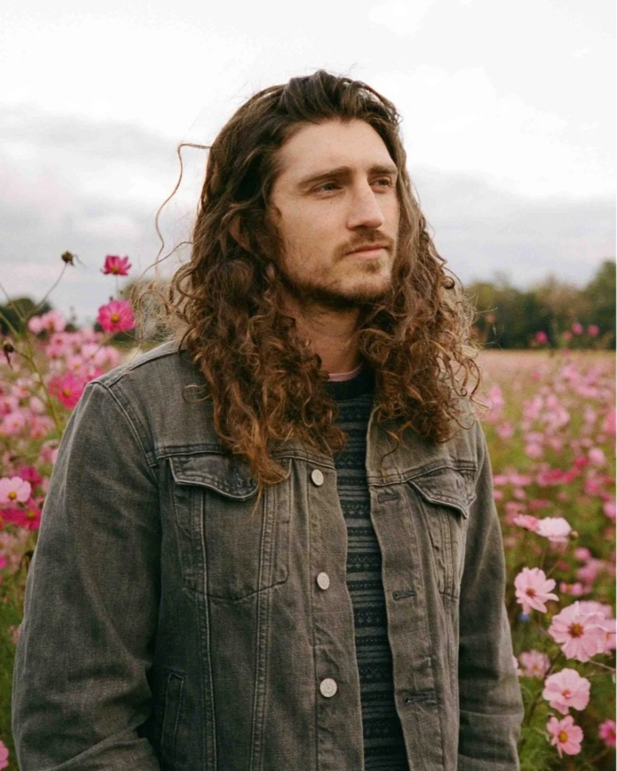 A young man with long curly hair and a beard stands in a field of pink flowers, wearing a denim jacket and a patterned shirt, with an overcast sky in the background.
