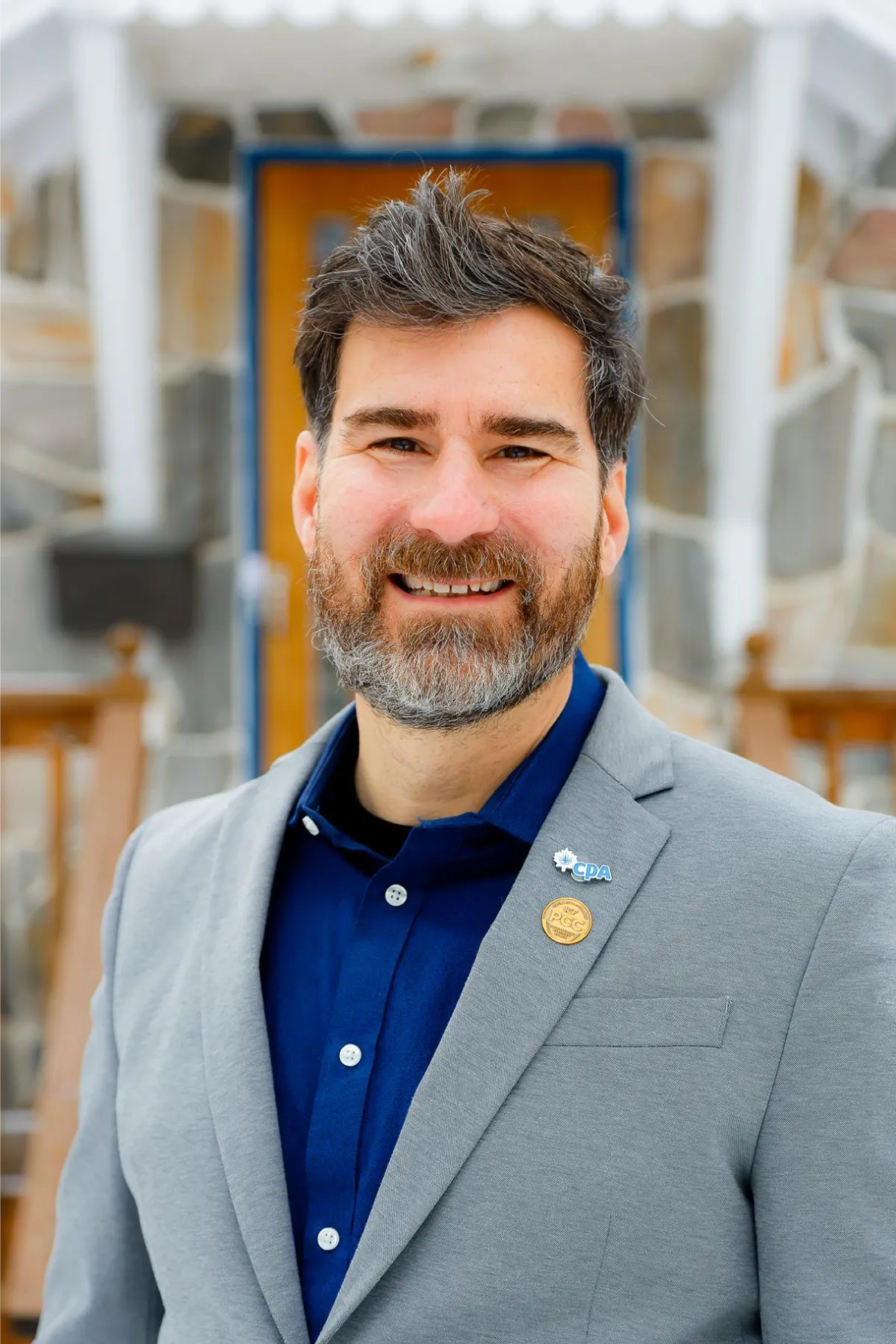 A smiling man with dark hair and a beard dressed in a gray blazer and navy blue shirt, standing in front of a lattice wood wall at an outdoor event.