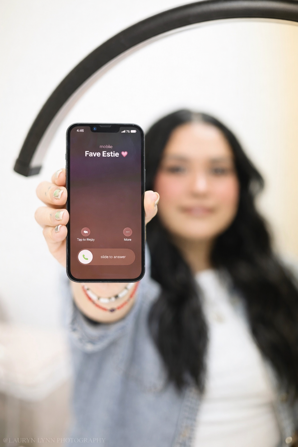 A young woman with long dark hair is holding a smartphone in front of her face showing an incoming call from a contact labeled "Fave Estie" with a pink heart emoji, blurred background, inside a room with a decorative mirror.