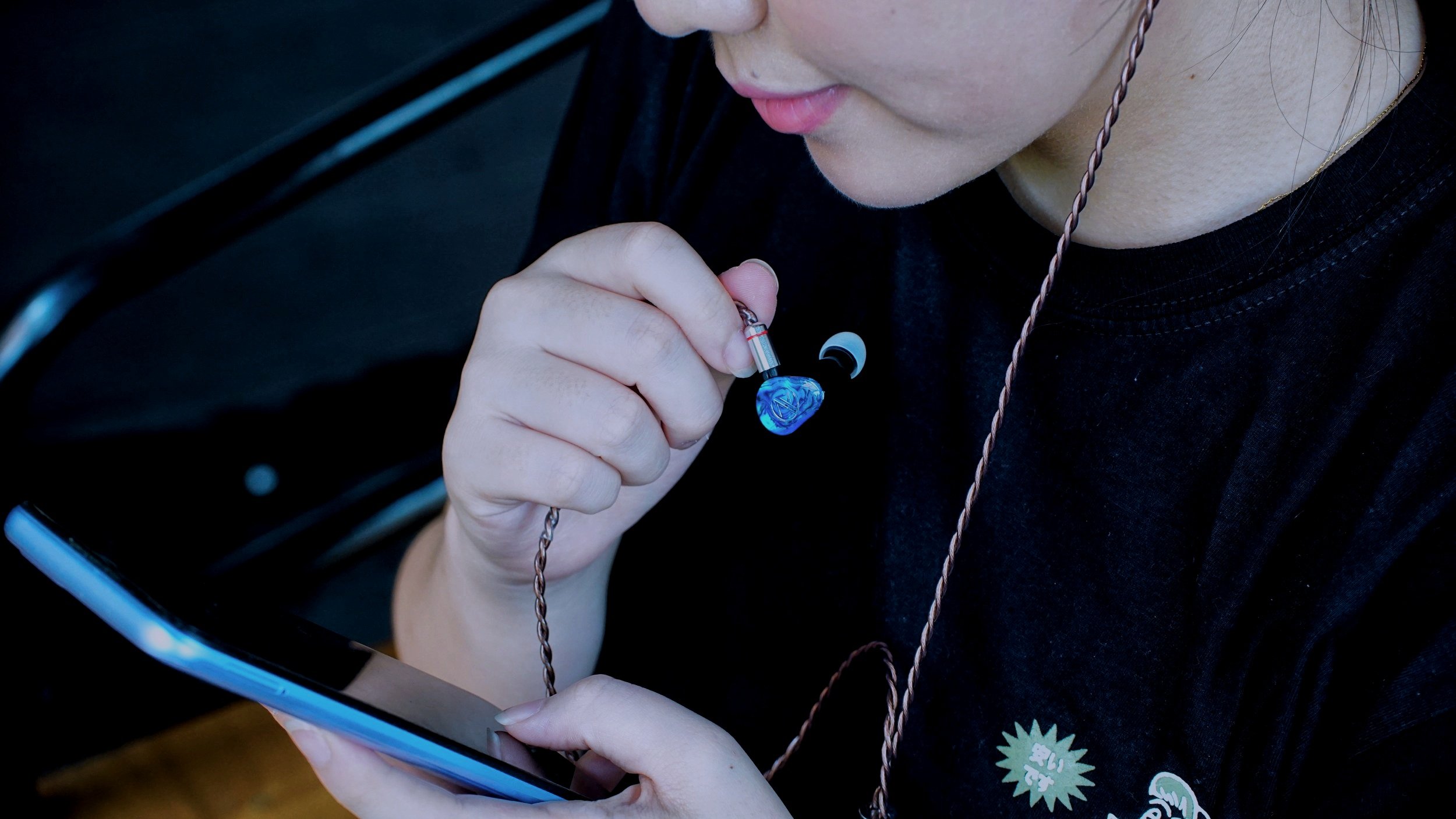 A person holding a colorful in-ear monitor while looking at a smartphone, with a black shirt and brown braided cable.