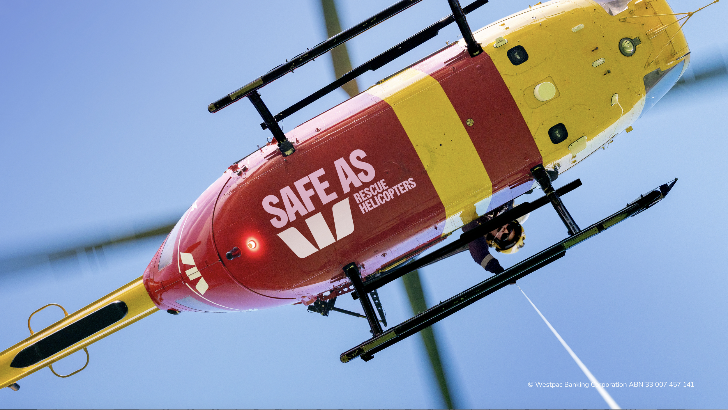 An rescue helicopter painted yellow and red with the words 'Safe as Rescue Helicopters' on its side, flying in clear blue sky.