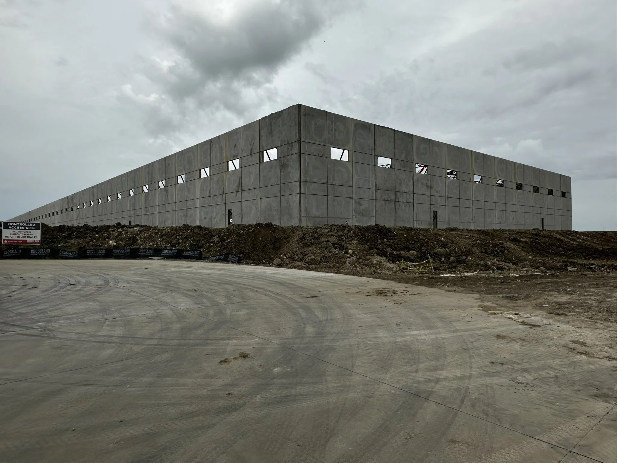 Under construction large concrete building with open windows, dirt and gravel in foreground, cloudy sky.