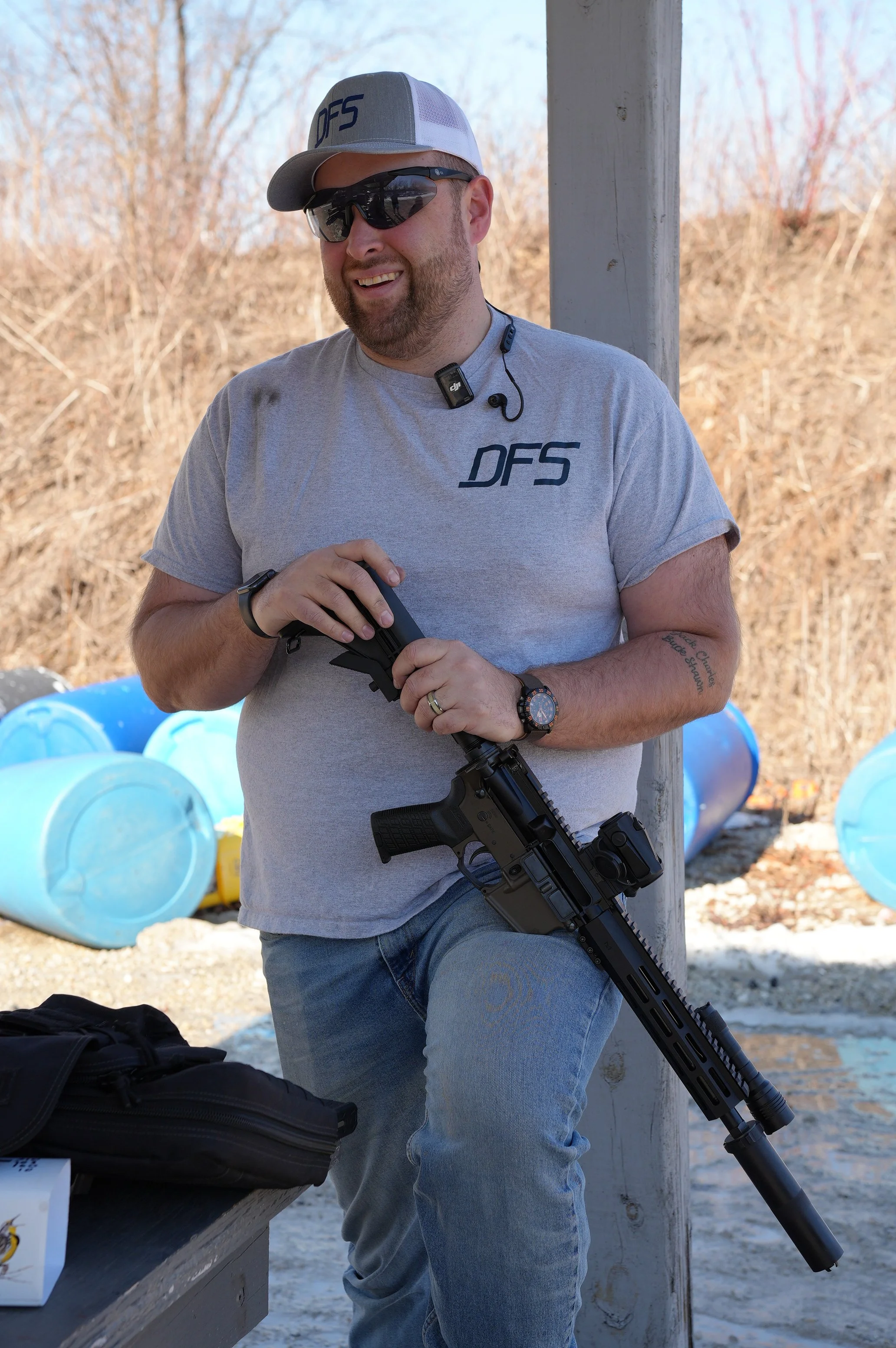A man with a beard, wearing sunglasses, a baseball cap, a gray DFS T-shirt, and jeans, is smiling while holding a semi-automatic rifle. He is standing outdoors near a wooden post, with blue barrels in the background and a table with a black bag on it.