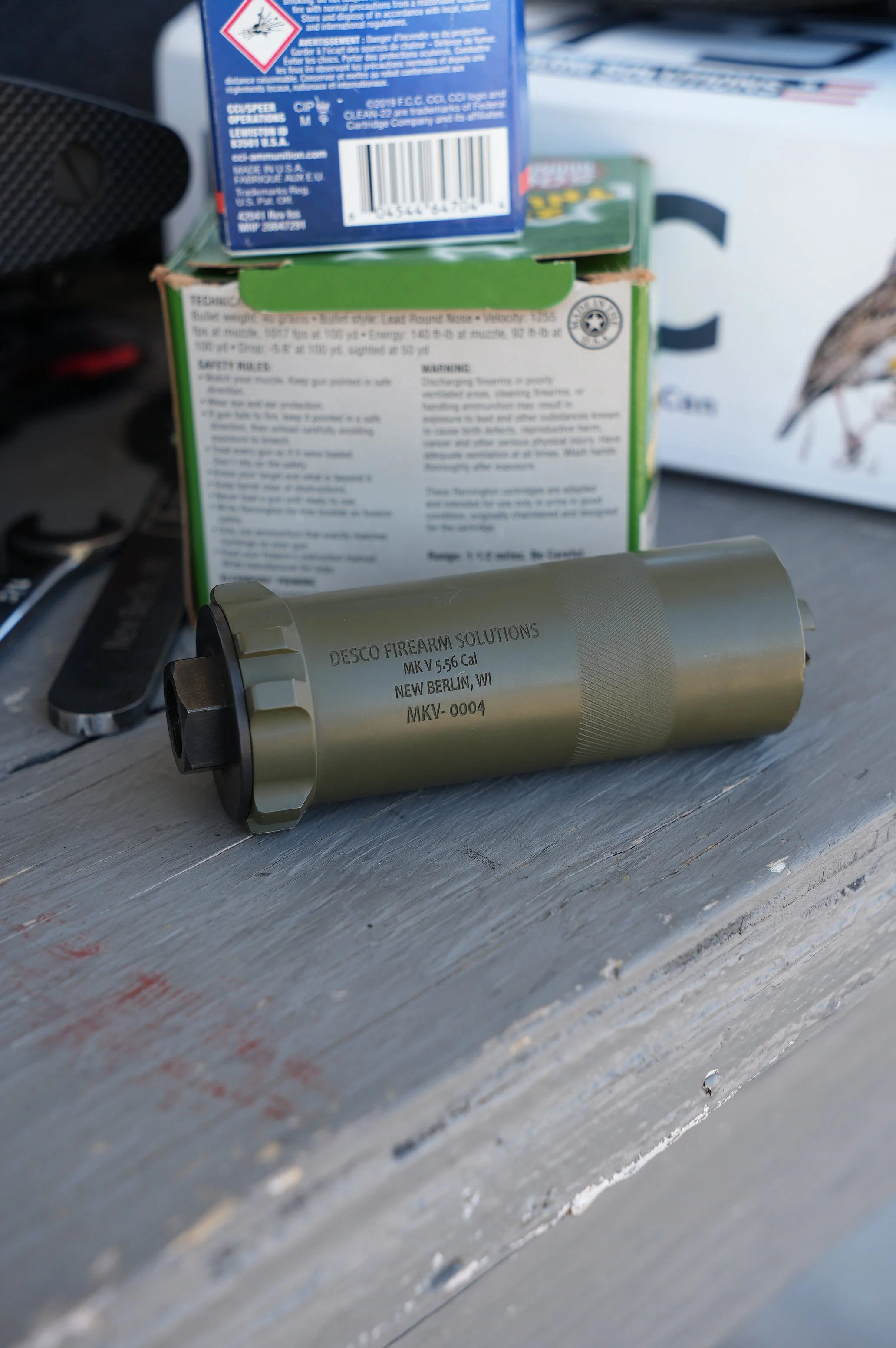 A cylindrical firearm suppressor with the inscription 'DESCO FIREARM SOLUTIONS MK V 5.56 Cal NEW BERLIN, WI MKV-0004' is placed on a weathered wooden surface. Behind it, there are ammunition boxes and a pocket knife.