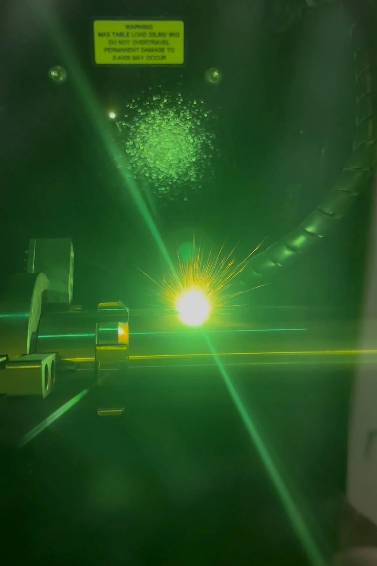 A close-up view of a laser welding process with a bright yellow-green laser beam and metal components, with condensation droplets on the surface and safety warning label above.