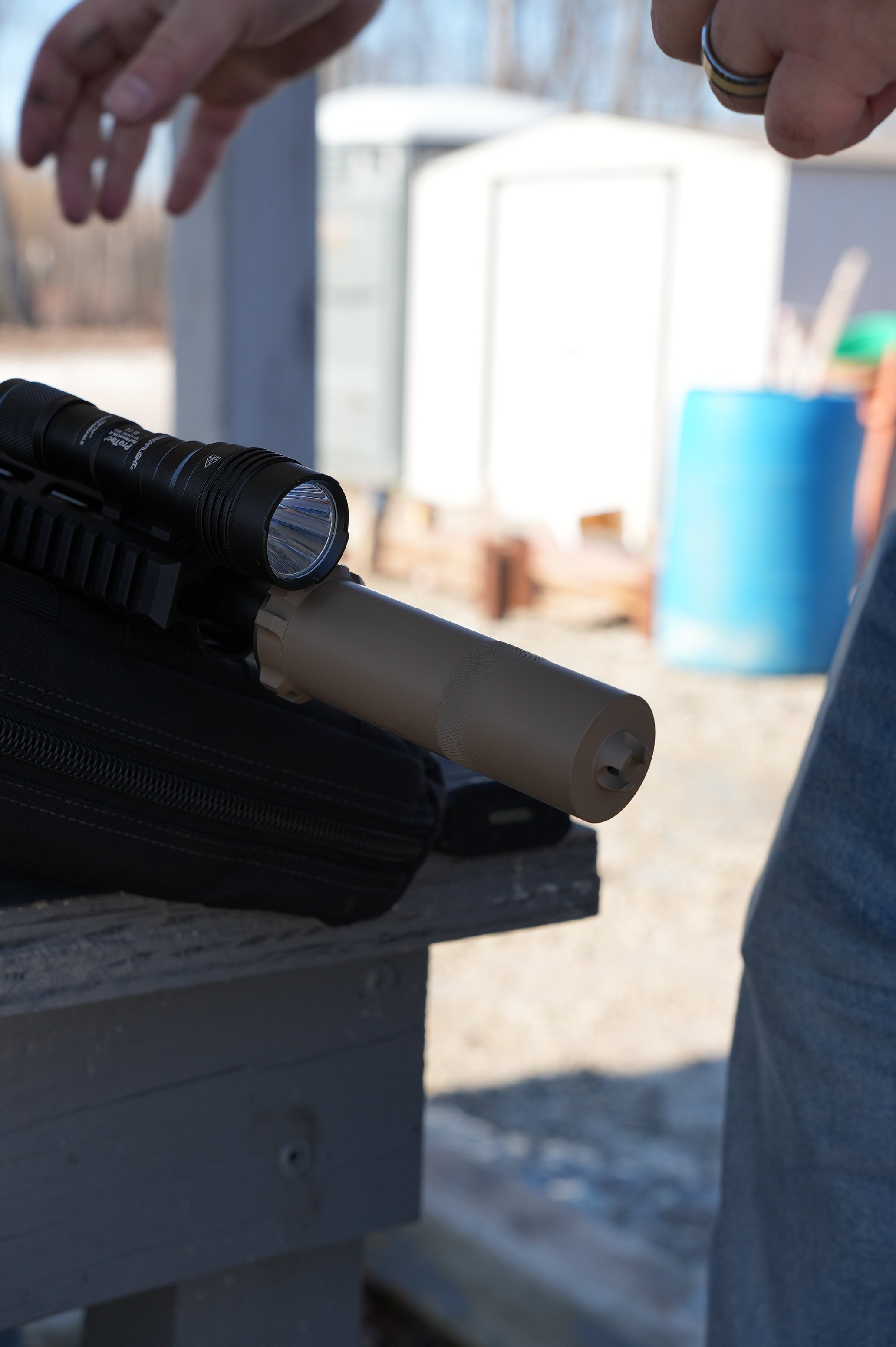 Close-up of a rifle with a scope and suppressor on a wooden surface, with someone adjusting or inspecting it.