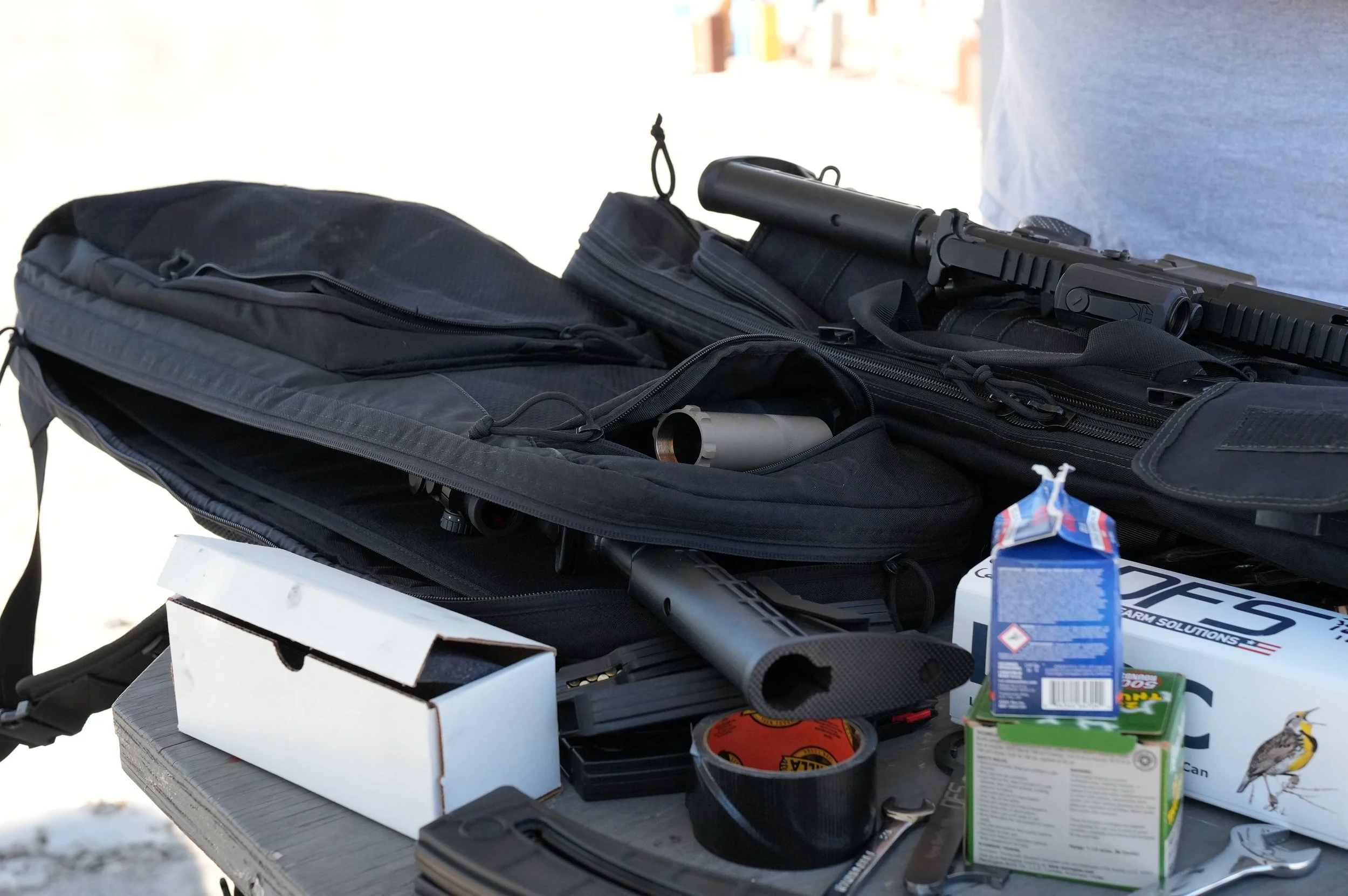 Table with a black gun, black tactical backpack, tissue box, duct tape, candy box, juice box, and small packaging.