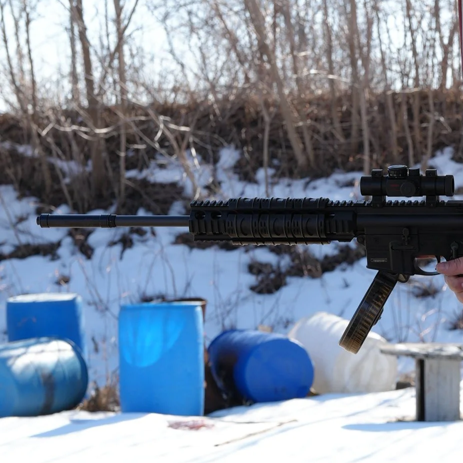 A person holding a black sniper rifle with a scope and suppressor, outdoors in a snowy area with blue barrels in the background.