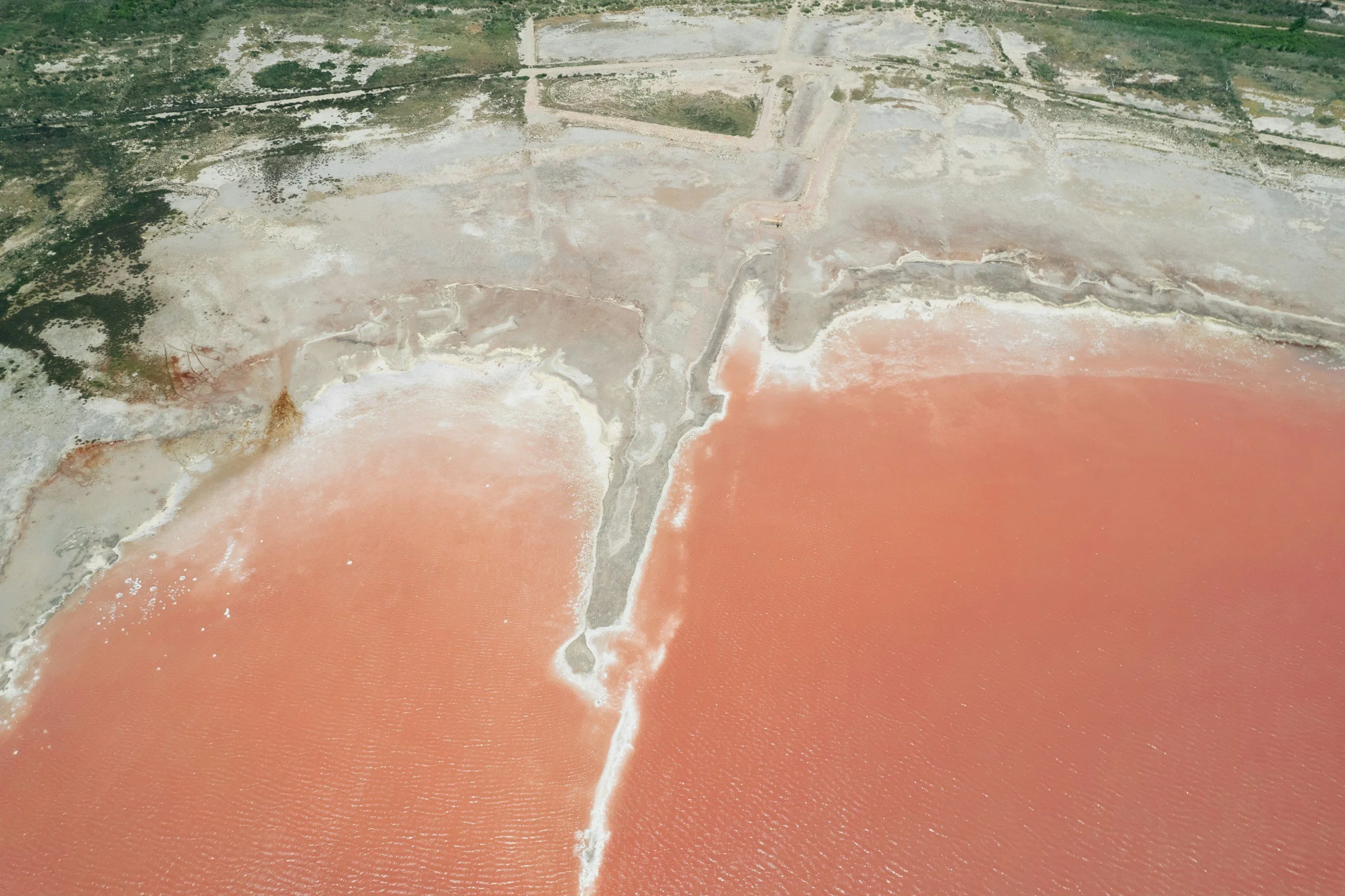 Aerial view of a coastal area with pink salt evaporation ponds separated by earthen embankments, alongside land with sparse vegetation.
