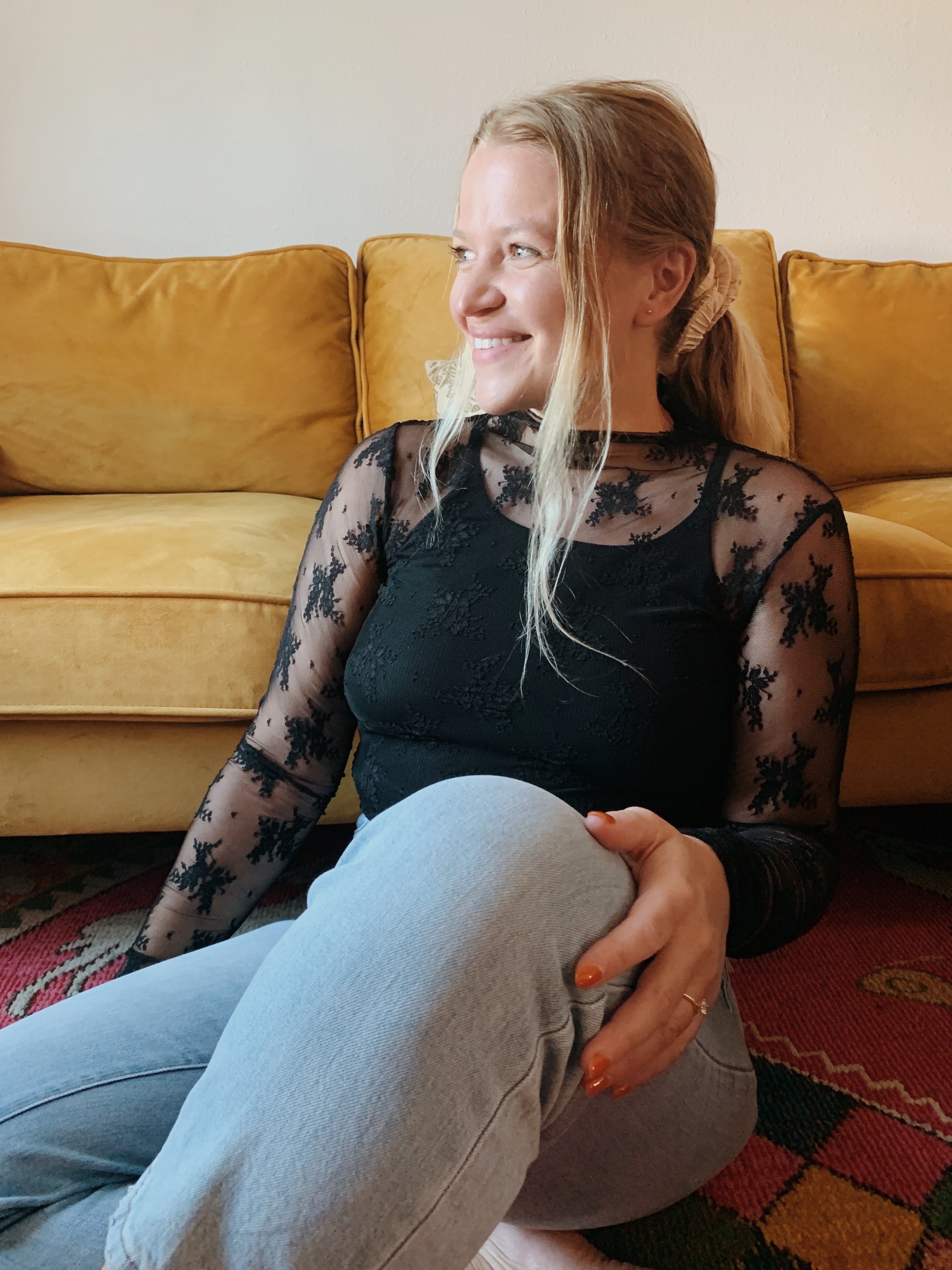 A woman with blonde hair sitting on a patterned rug in front of a yellow sofa, smiling and looking to her right, wearing a black lace top and light gray pants.