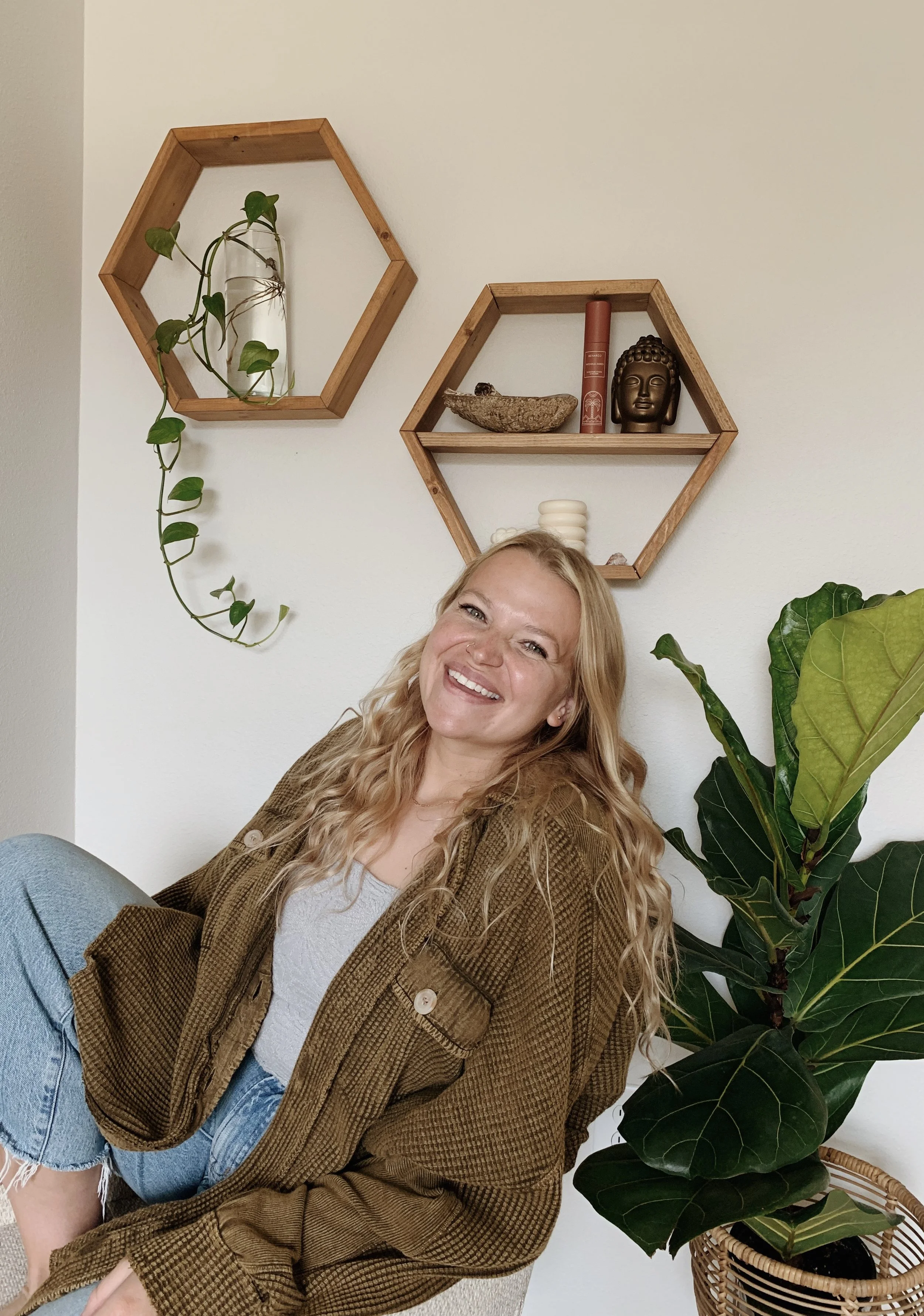 A woman with long blonde curly hair, smiling and sitting near a large green plant, with decorative wall shelves behind her containing various objects including a buddha head, books, and decorative items.