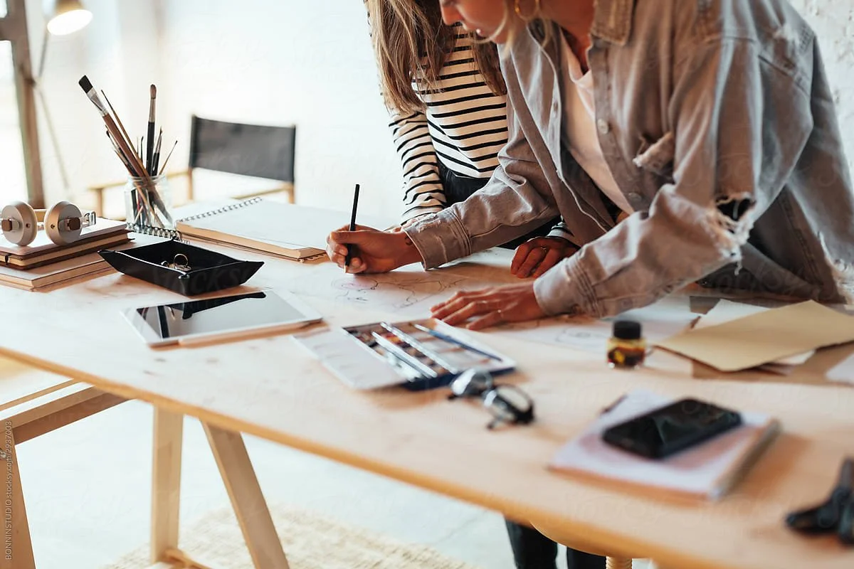 two people working a drafting desk with art supplies