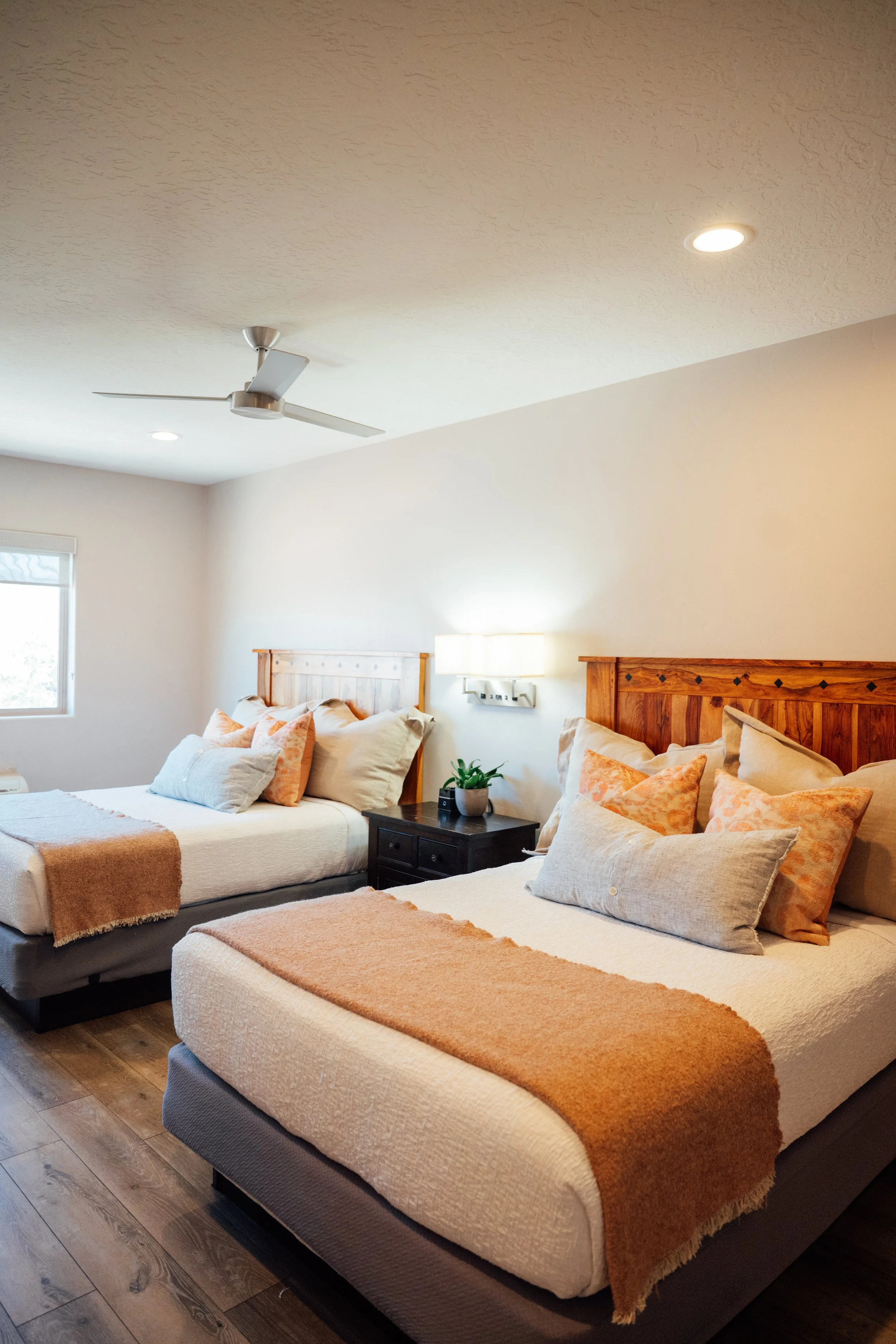 Movara double queen room with beige and peach bedding, multiple pillows, wooden headboards, bedside table with a small plant, and a ceiling fan in a bedroom with hardwood floors and a window.