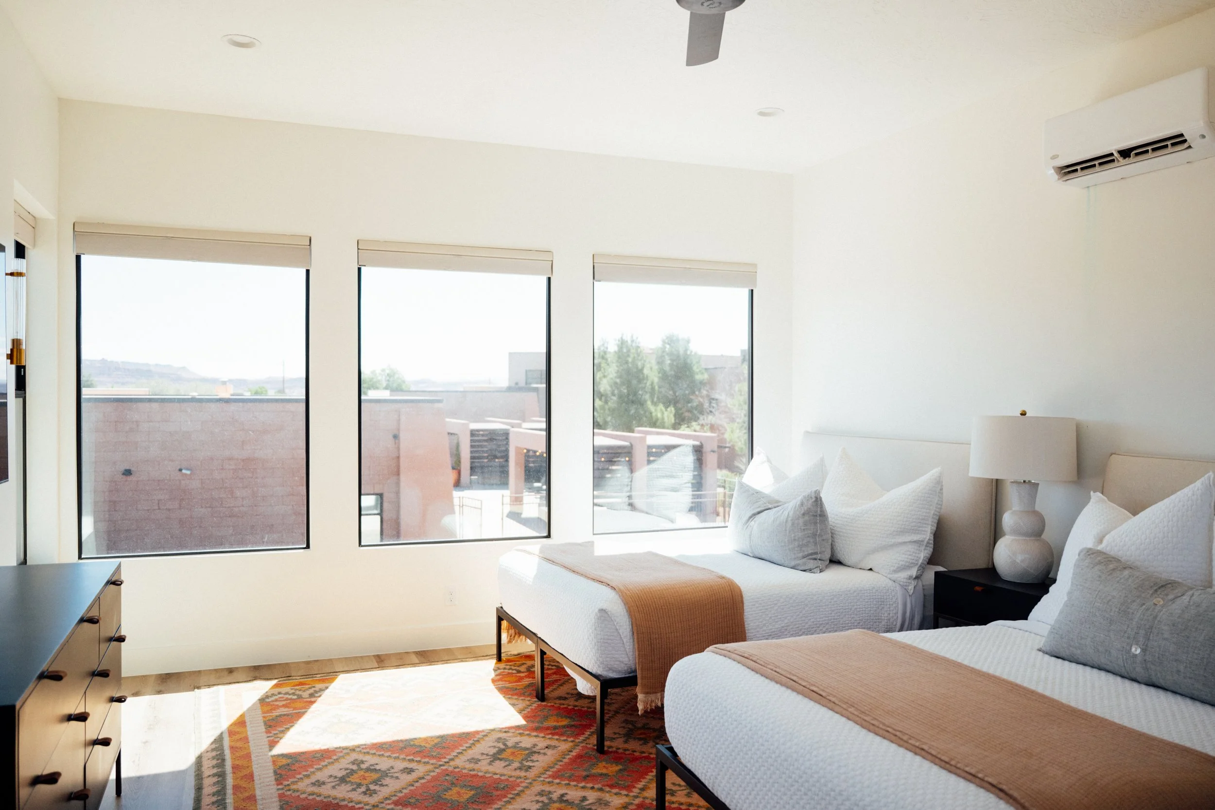 Movara Villa double queen room with large windows showing an outdoor view, a bed with white bedding and a tan blanket, a white lamp on a black nightstand, a wooden dresser, and a colorful patterned rug.