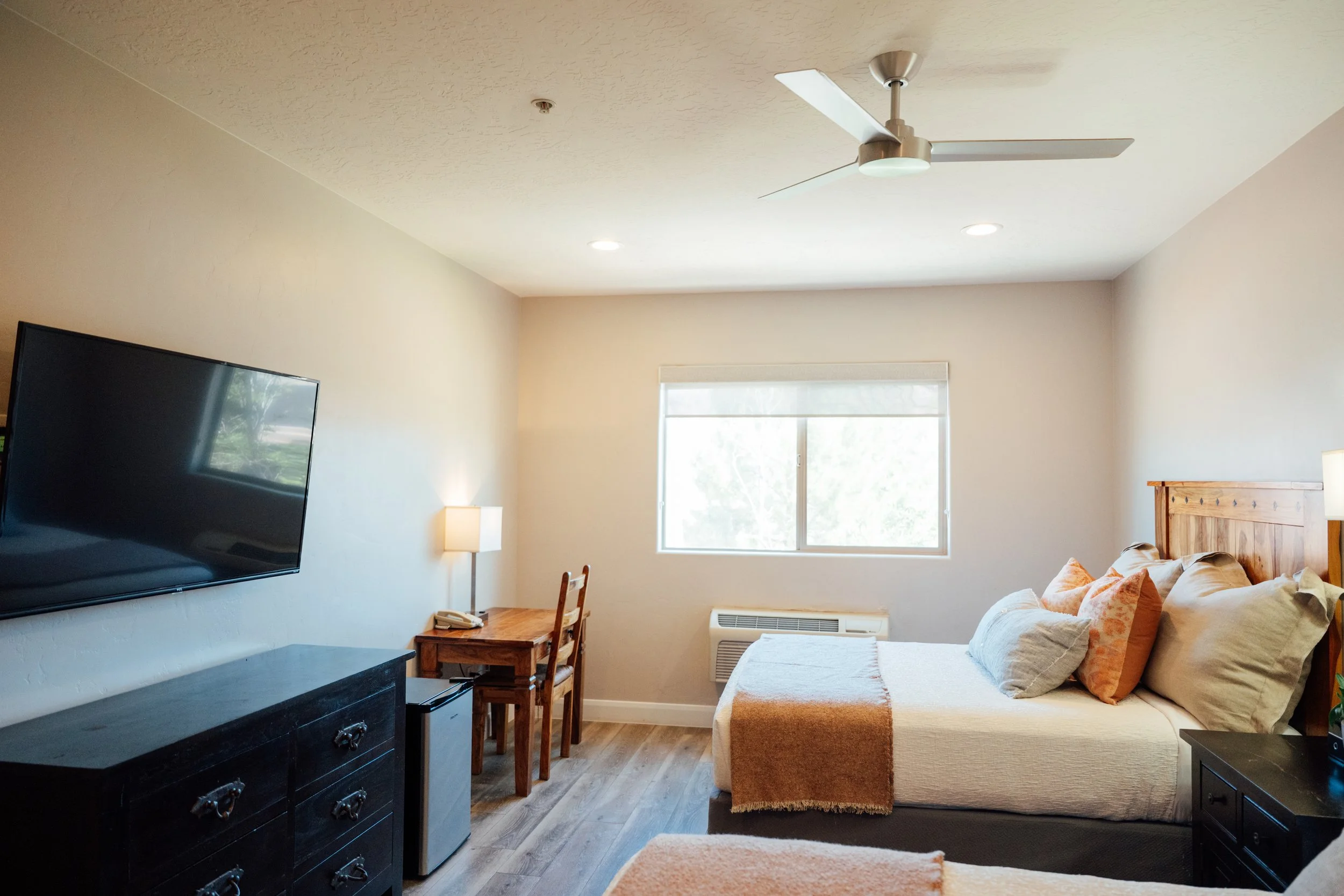 Movara double queen room with a wall-mounted flat-screen TV, a wooden table with a telephone and chairs, a window with blinds, a ceiling fan, a sofa with cushions, a lamp, and wooden flooring.