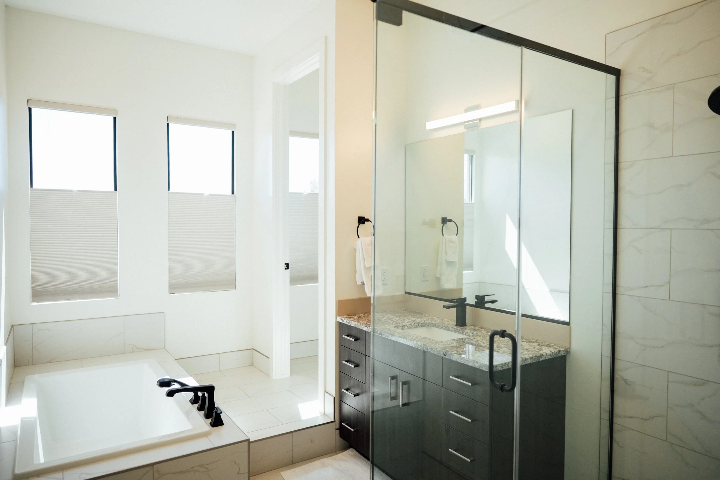 Modern bathroom featuring a large mirror, black faucets, a stone countertop with drawers, a glass shower door, and two tall windows with blinds, with a bathtub visible in the foreground.