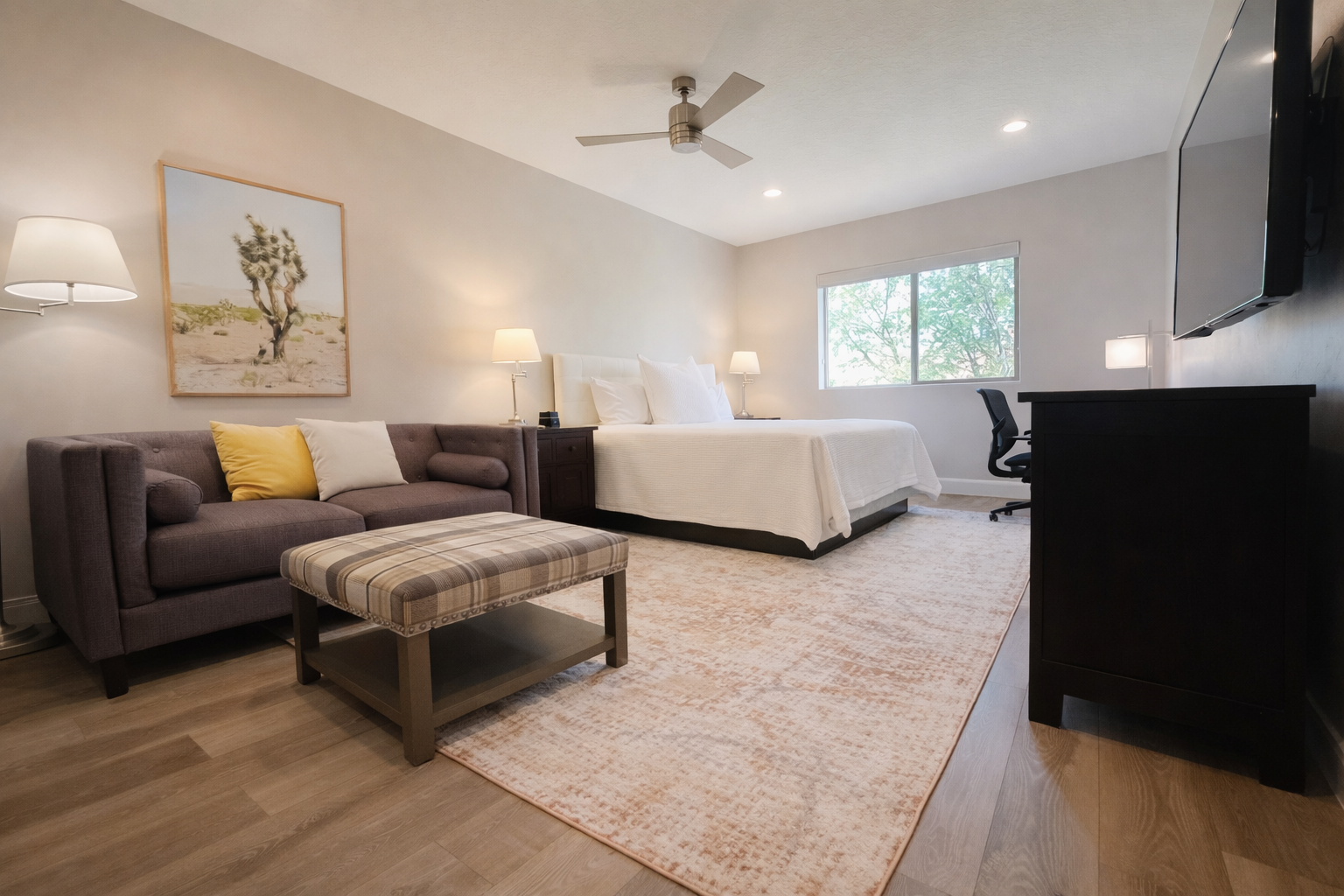 Modern bedroom with a bed, dark sofa, plaid ottoman, ceiling fan, and framed desert artwork. There are multiple lamps, a large window with natural light, and a desk with a chair.