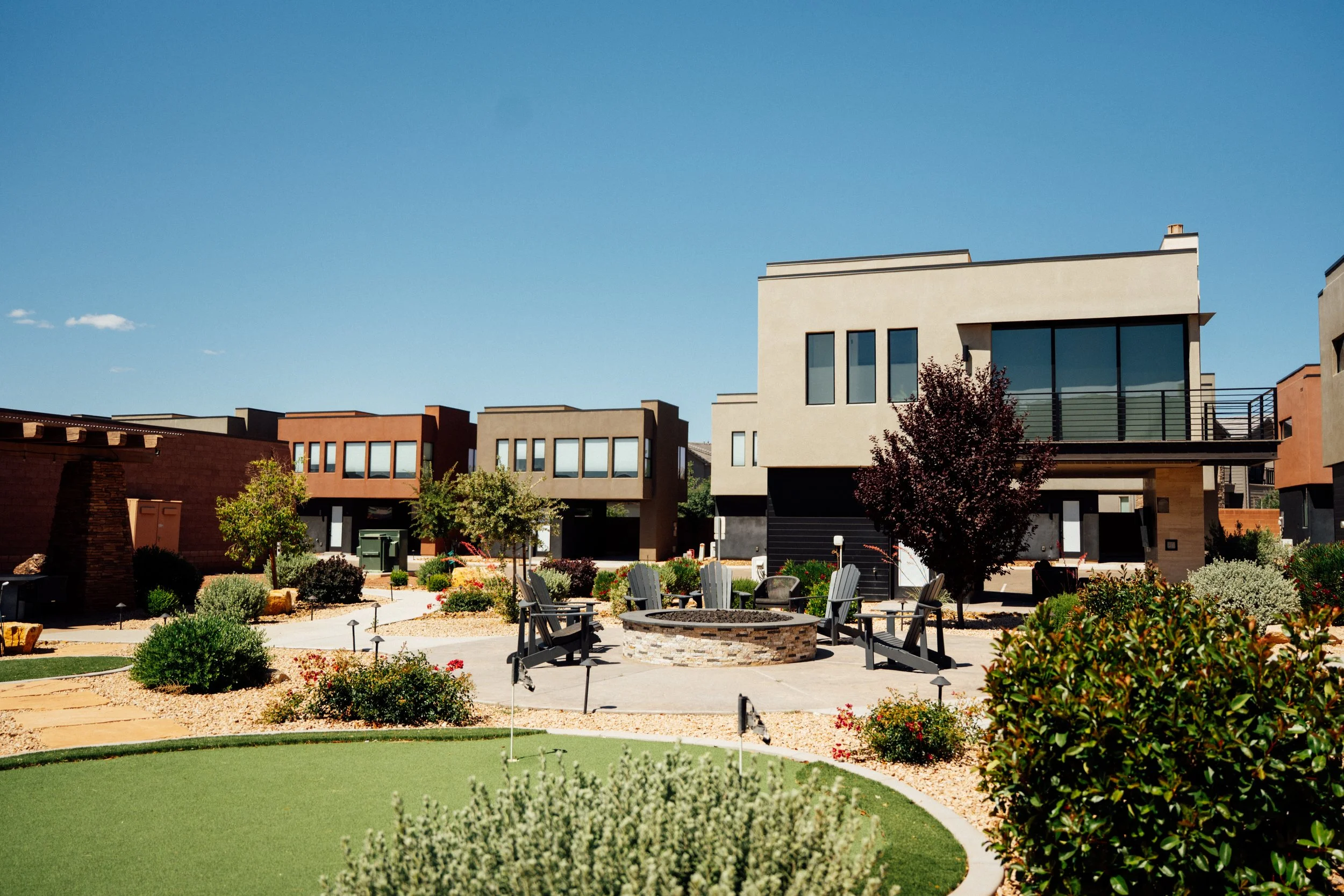 A backyard with a putting green, outdoor furniture, and landscaped garden with modern houses in the background under a clear blue sky.