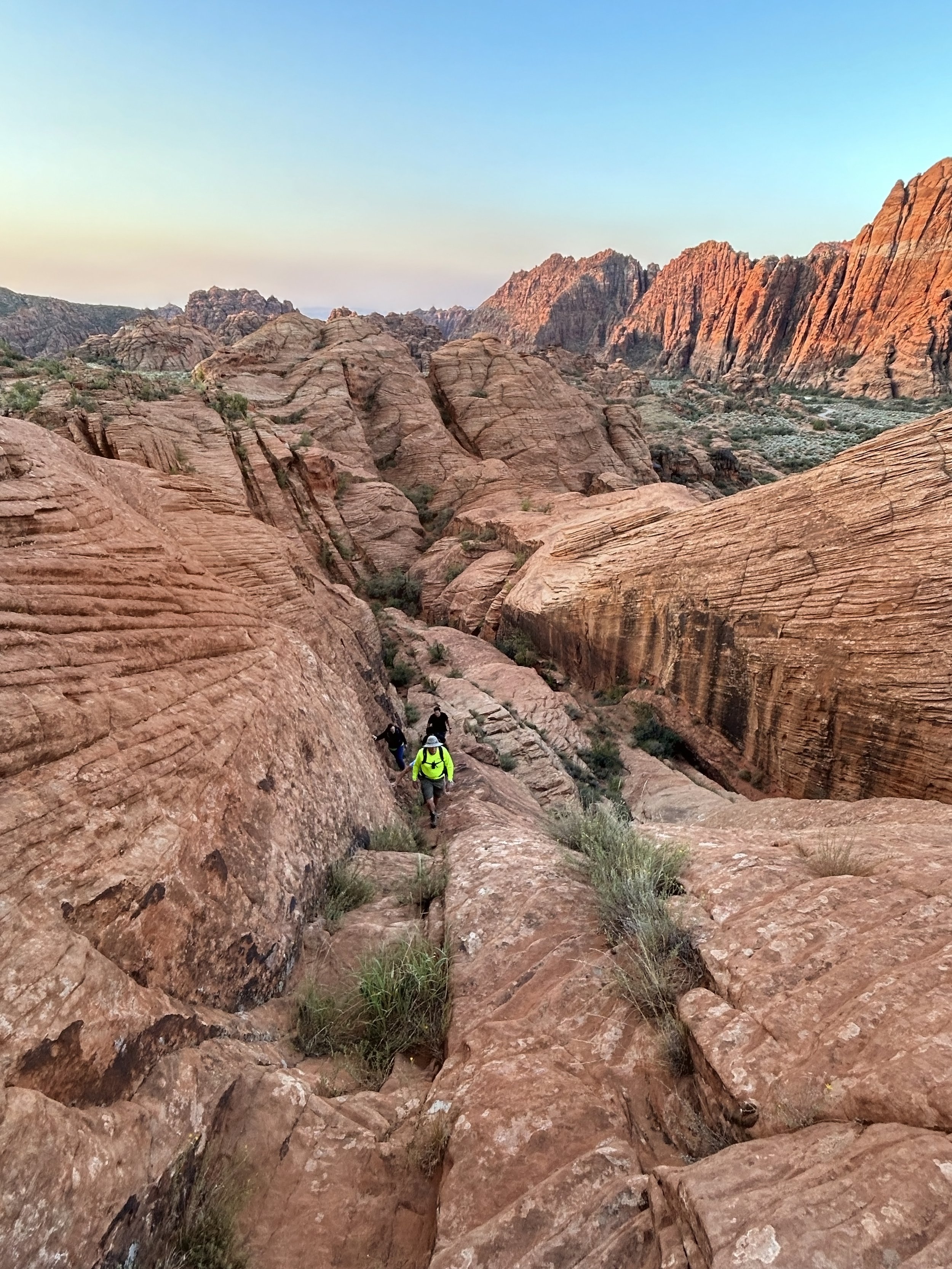 Hikers climbing a rugged red rock trail in a desert canyon with towering sandstone formations at sunset.