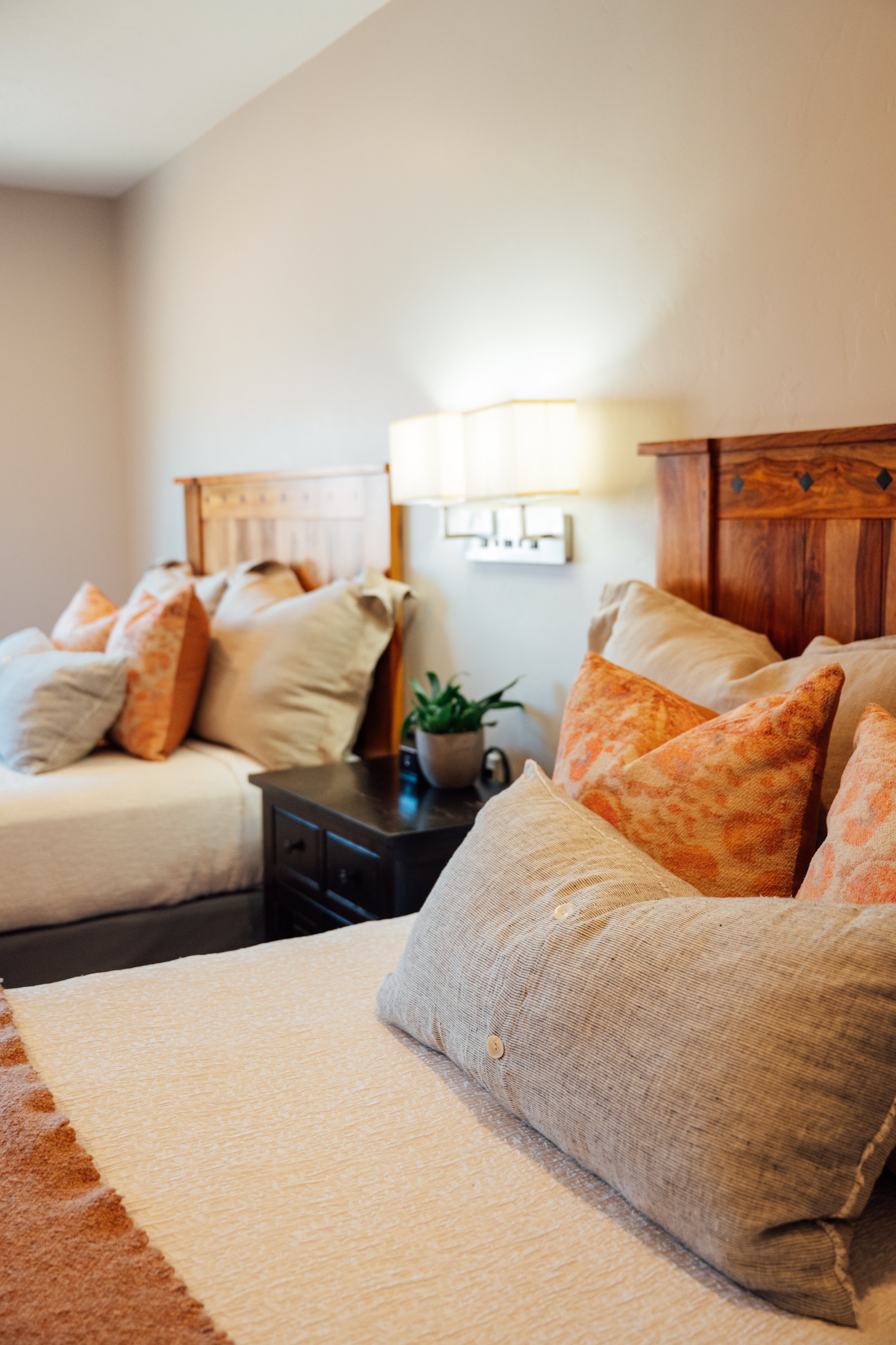 Movara double queen room with two beds, each with beige and orange pillows, a wooden headboard, a black nightstand with a potted plant, a wall-mounted light fixture, and neutral-colored walls.