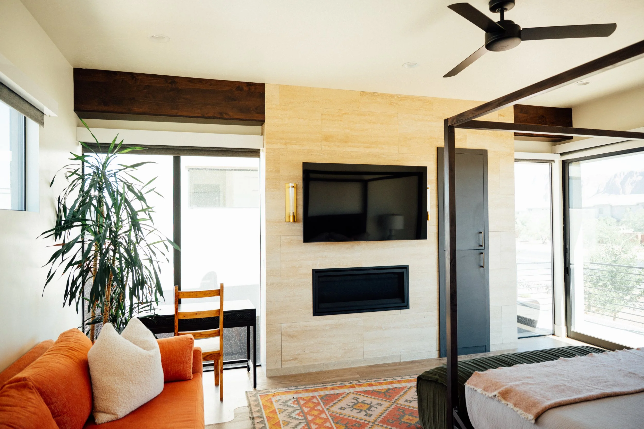 Living room with an orange sofa, decorative pillows, a large potted plant, a wall-mounted TV over a fireplace, hardwood and tile flooring, large windows with view, and a ceiling fan.
