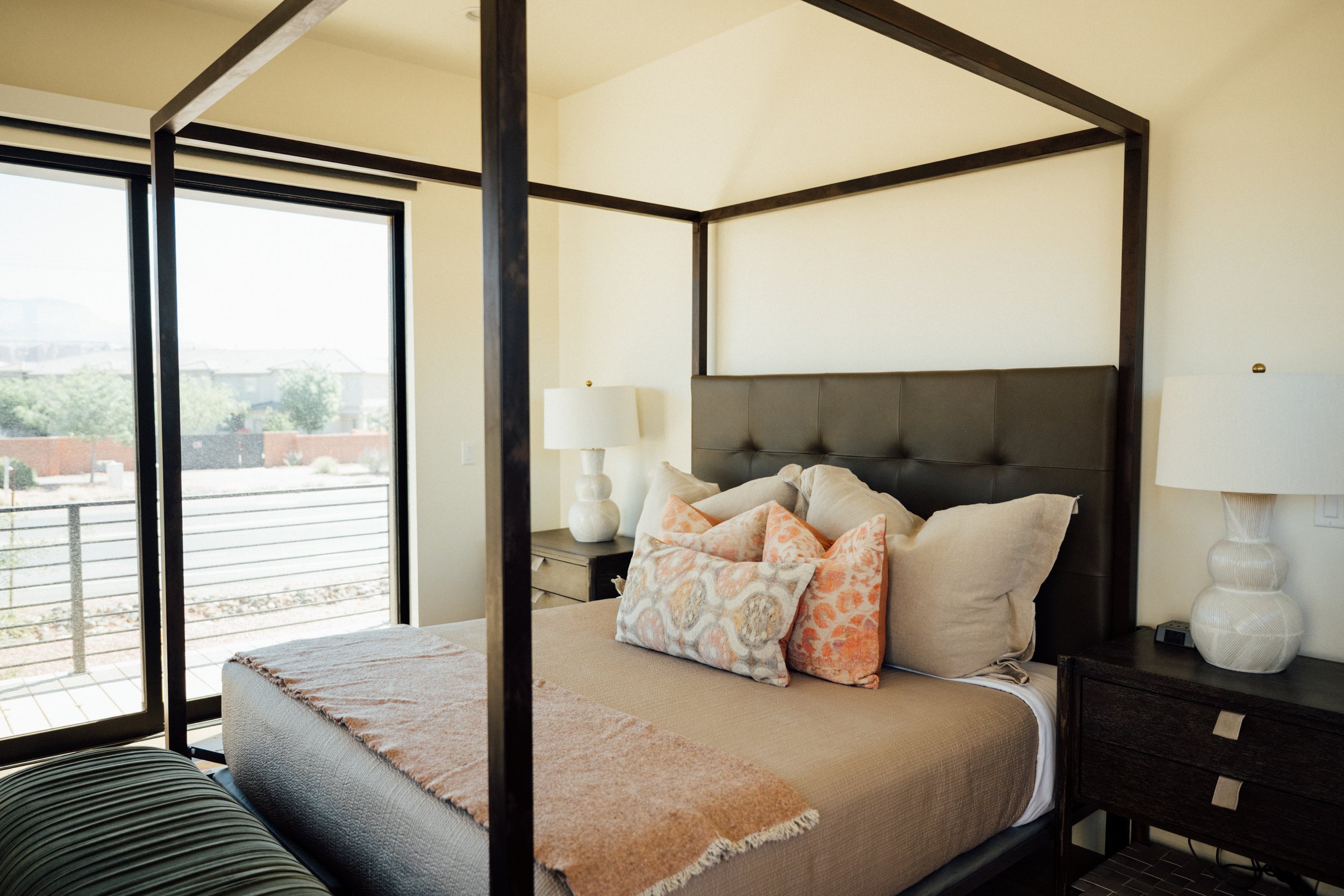 Movara Villa primary suite with a large bed covered in beige and orange pillows, flanked by white nightstands with matching lamps, large windows showing an outdoor view, and a minimalist interior design with dark wood accents.