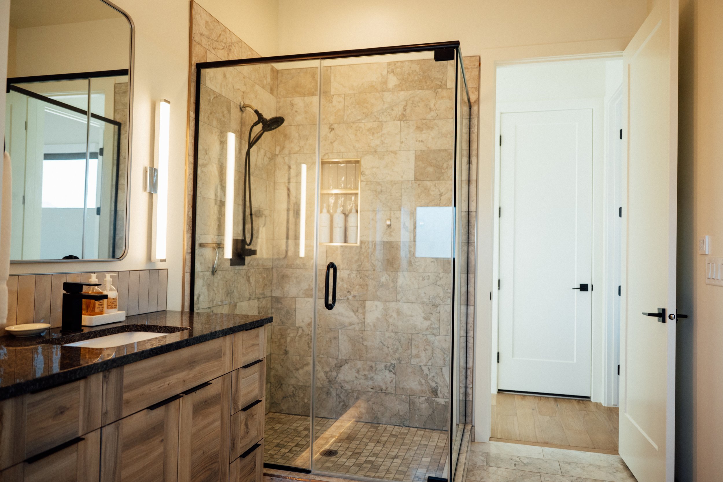 A bathroom with a walk-in shower enclosed with glass, tiled in beige marble, with a black shower head and a shower niche. There is a wooden vanity with a dark countertop and a large mirror above, and a white door in the background.