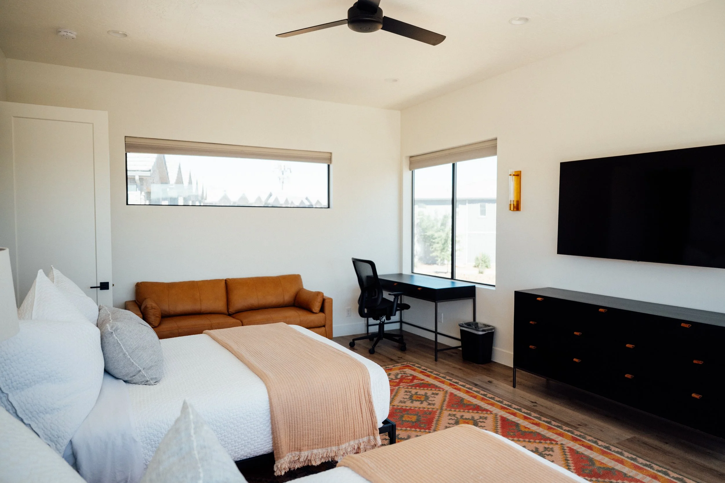 A modern living room with a white bed in the foreground, an orange sofa against the wall, large windows allowing natural light, a black desk with an office chair, and a flat-screen TV mounted on the wall. The room features a ceiling fan and a colorfu