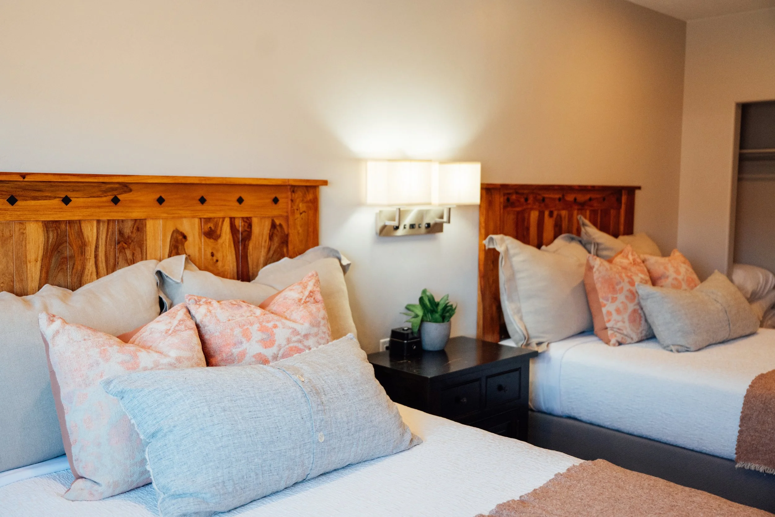 Movara double queen room with two beds with wooden headboards and multiple pillows, a nightstand with a plant and alarm clock, wall lamp, and closet in the bedroom.