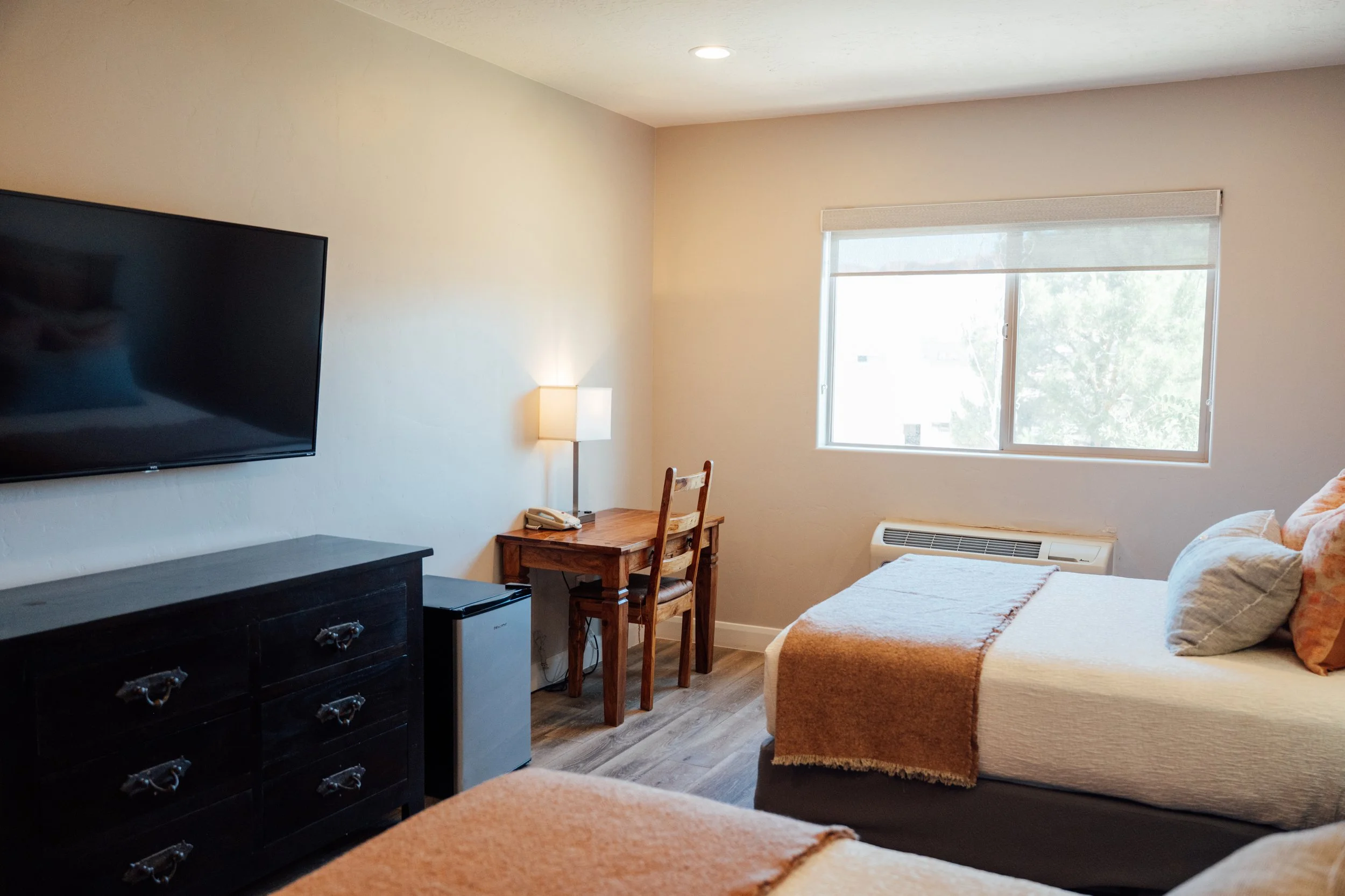 Movara double queen room with a large window, a flat-screen TV mounted on the wall, a wooden desk with a chair, a small refrigerator, a lamp, and a bed with an orange blanket and pillows.