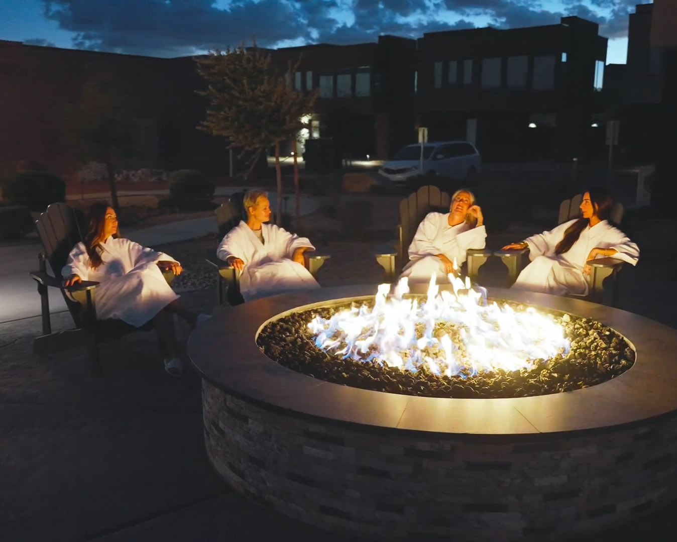 Four women sitting around a lit outdoor fire pit at night, all dressed in white robes, in a relaxed setting