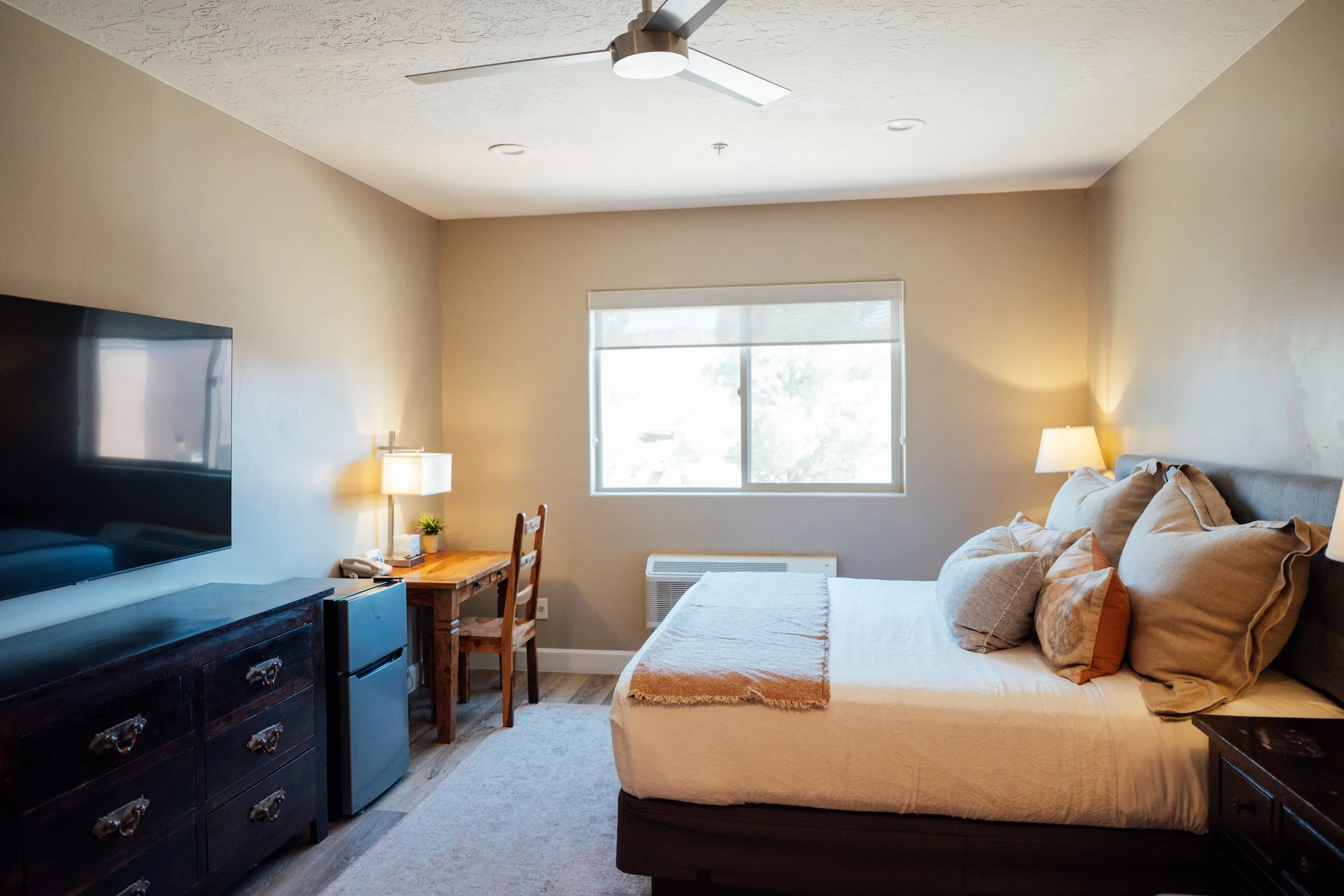 Movara single king room with A cozy living room with a large window, beige walls, a bed with pillows, a wooden desk with a lamp and plant, a TV, a ceiling fan, and wooden flooring.