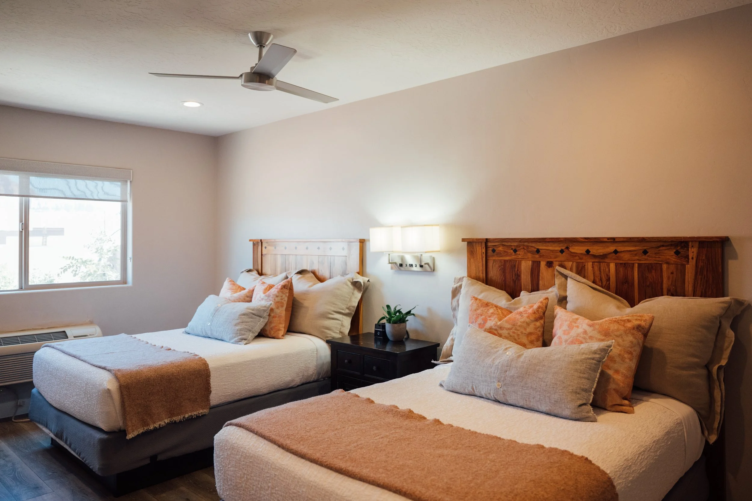 Movara Double Queen room with two beds, wooden headboards, multiple pillows, a small black nightstand with a potted plant, wall-mounted light fixture, window with blinds, ceiling fan, and beige walls.