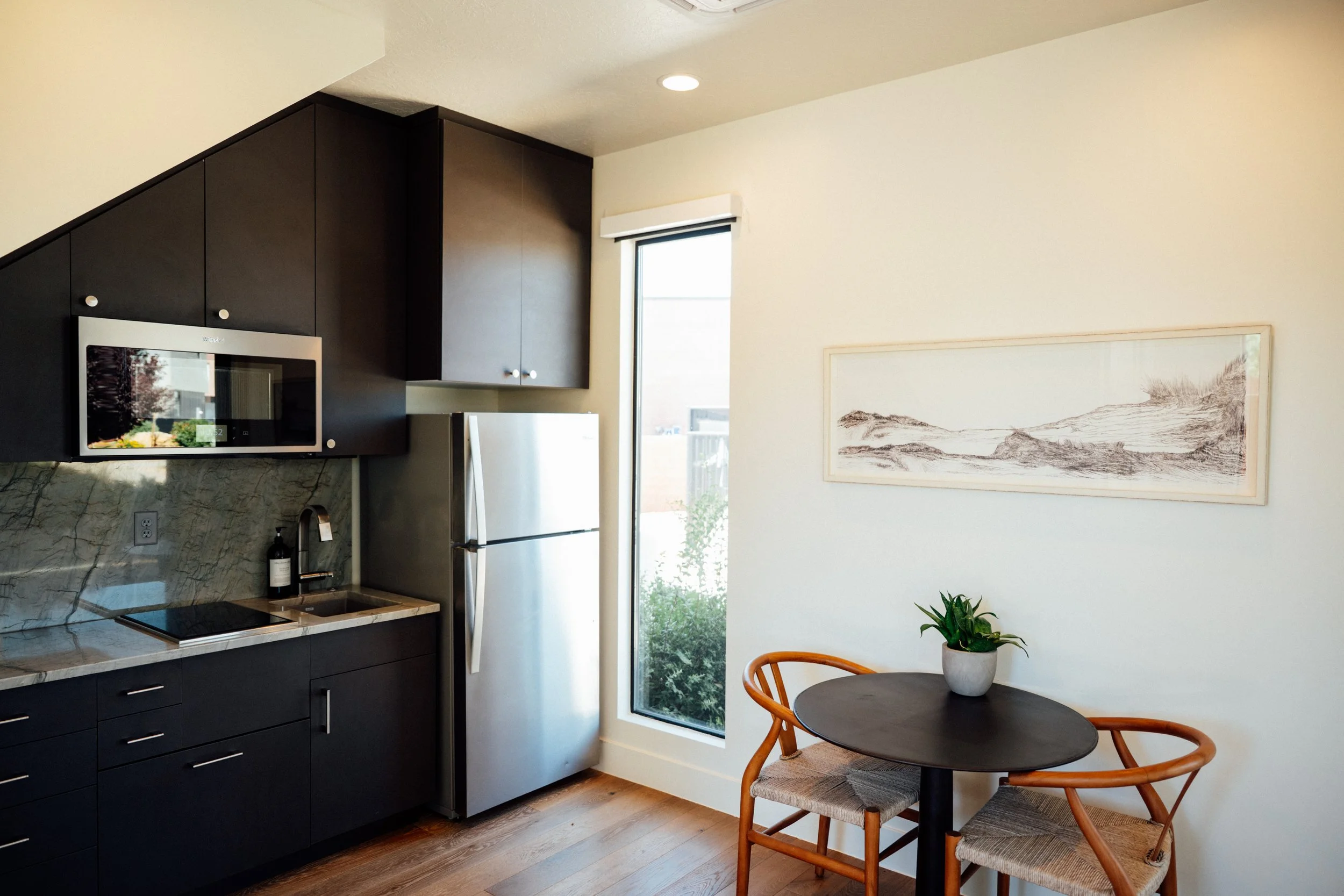 A modern kitchen with dark cabinets, a stainless steel microwave, a white refrigerator, a marble countertop, and a small round dining table with two wooden chairs and a potted plant. There's a large window with a view outside and a framed landscape p