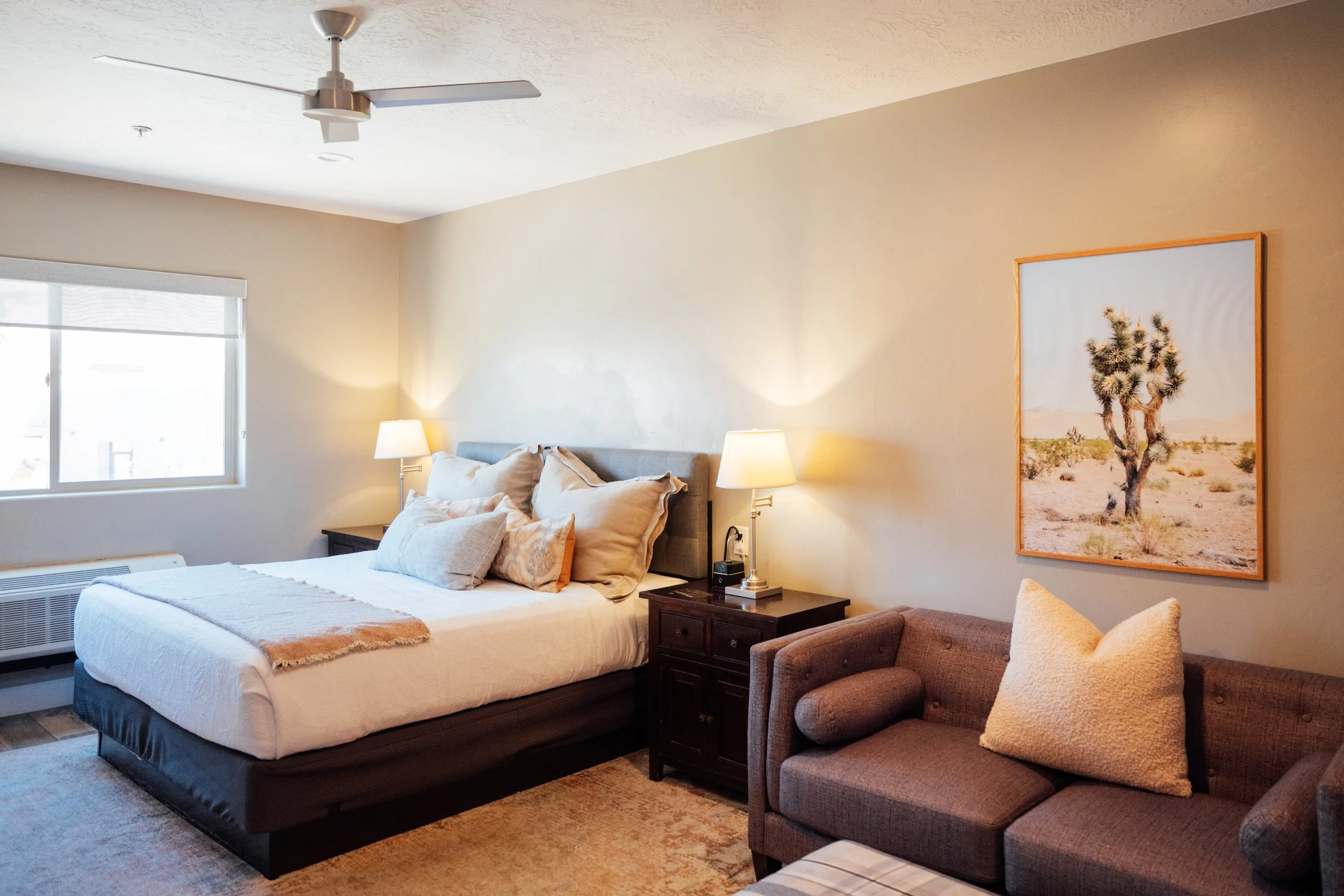 A bedroom with a bed, pillows, a bedside table with lamps, a framed desert picture, a window, and a ceiling fan.