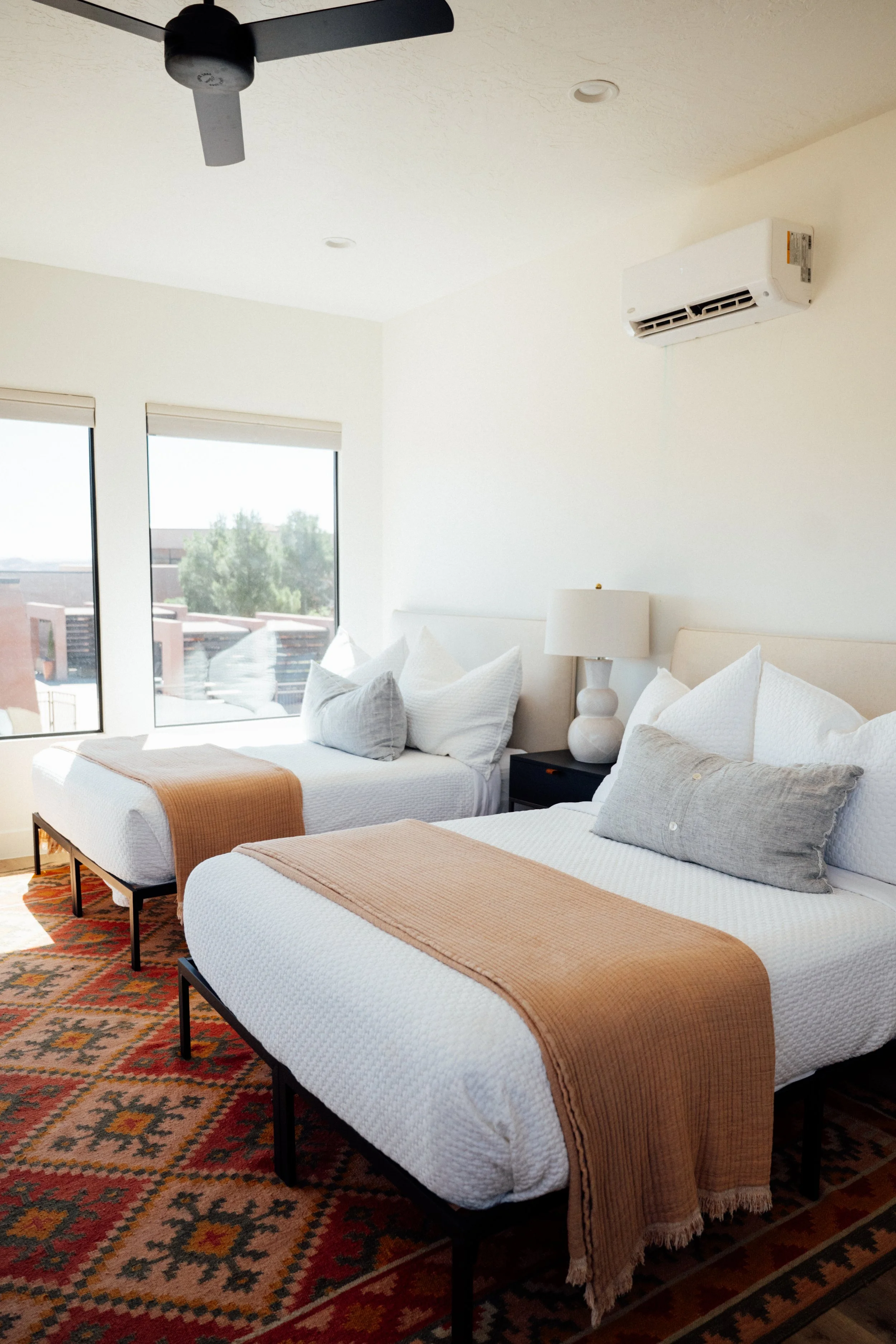 Movara double queen villa with two beds with beige throw blankets, pillows, and white quilted covers in a bright room with large windows, a nightstand with a lamp, ceiling fan, and a wall-mounted air conditioning unit.