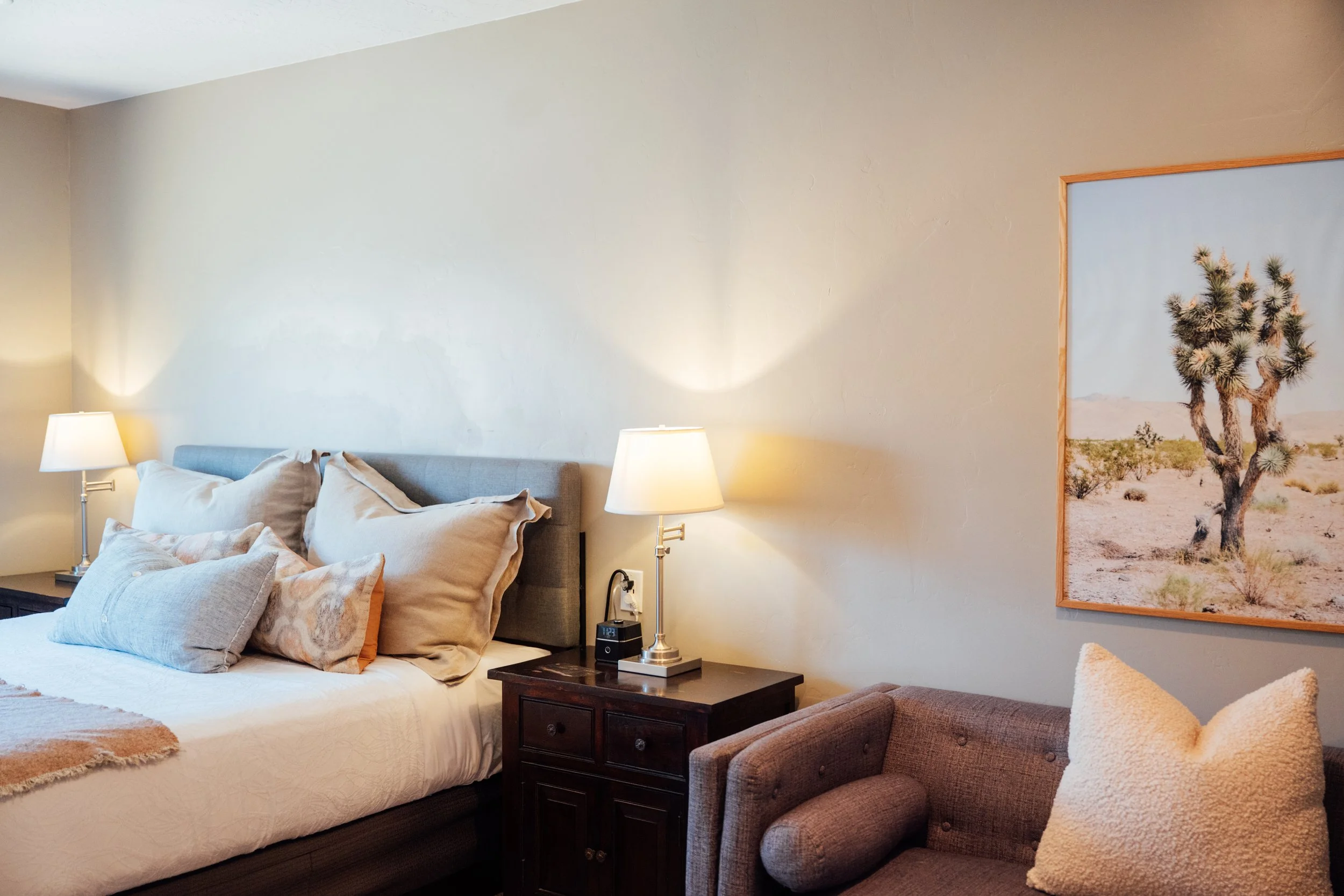 A bedroom with a bed, pillows, two bedside lamps, a wooden nightstand, a framed desert scene with a cactus, and a gray armchair with a textured pillow.