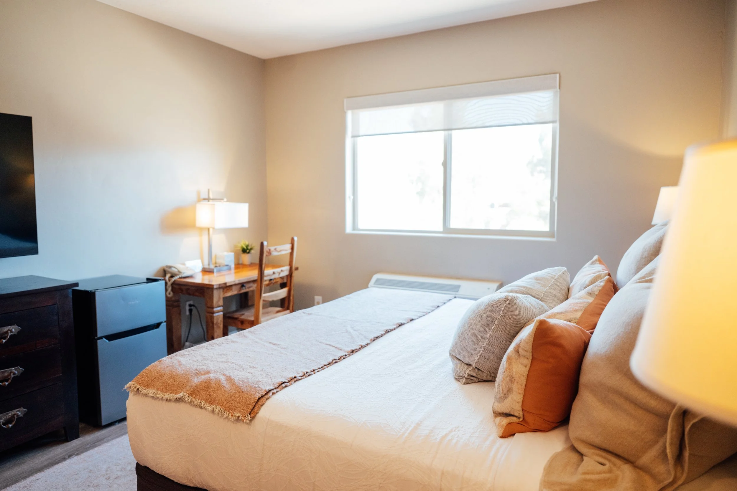 Movara single king bed room with beige bedding, and several pillows. There is a window with sunlight shining through. A small wooden desk with a lamp, some potted plants, and miscellaneous items is beside the bed. 