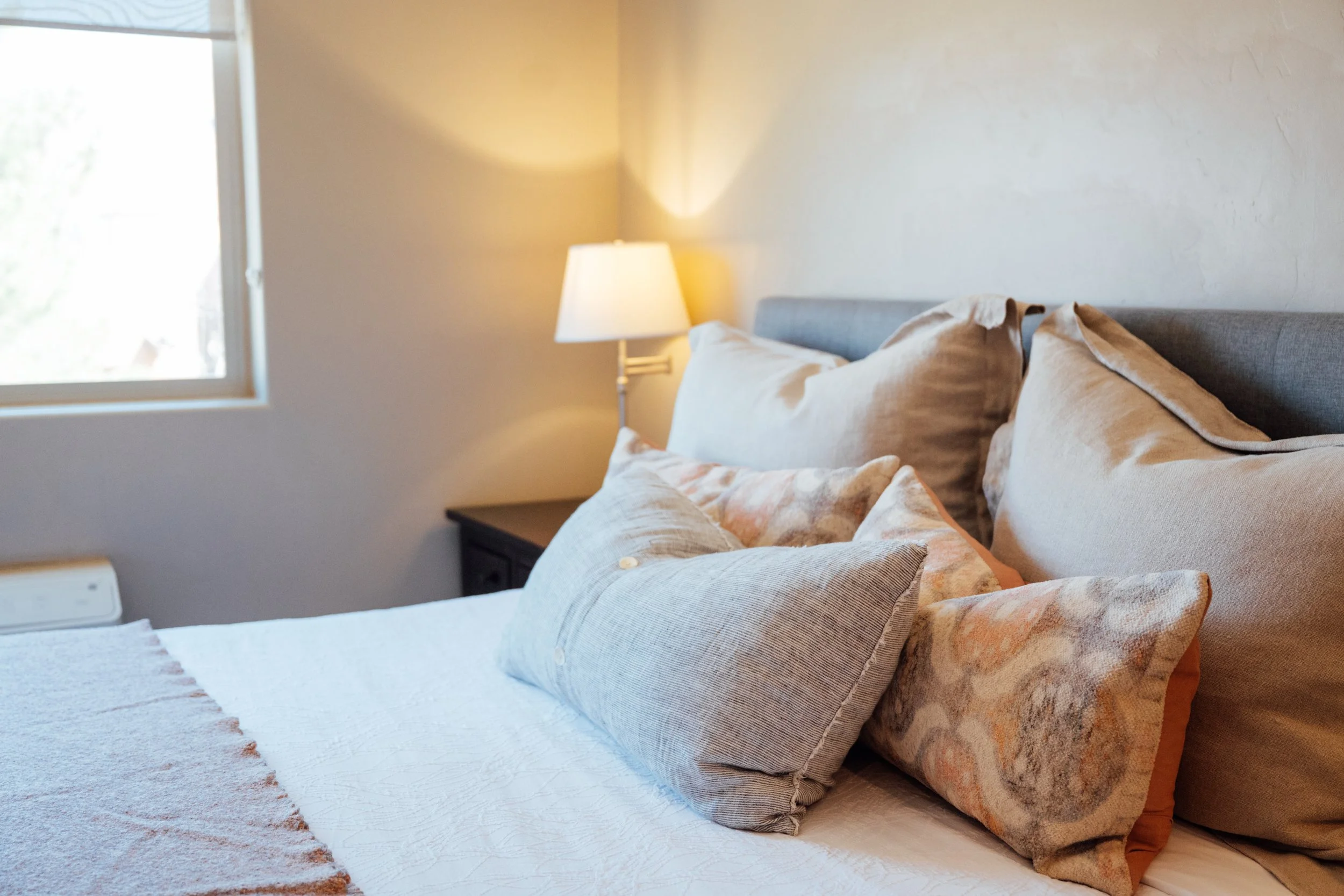 Movara single king bed room with A neatly made bed with beige pillows and a textured white bedspread in a cozy bedroom, illuminated by a table lamp on the bedside table, with a window letting in natural light.