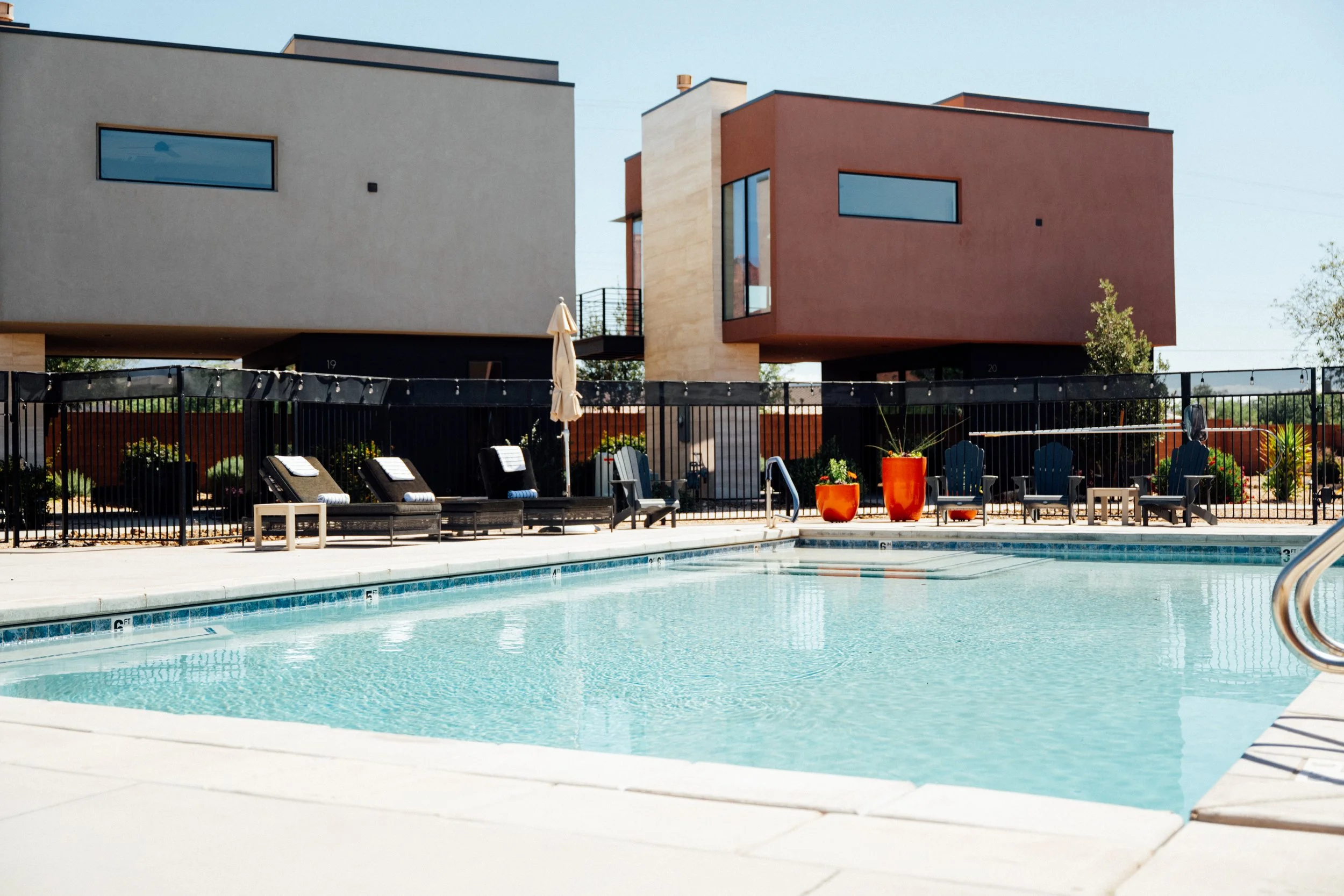 An outdoor swimming pool area with lounge chairs and large orange flower pots in front of modern residential buildings with geometric design.