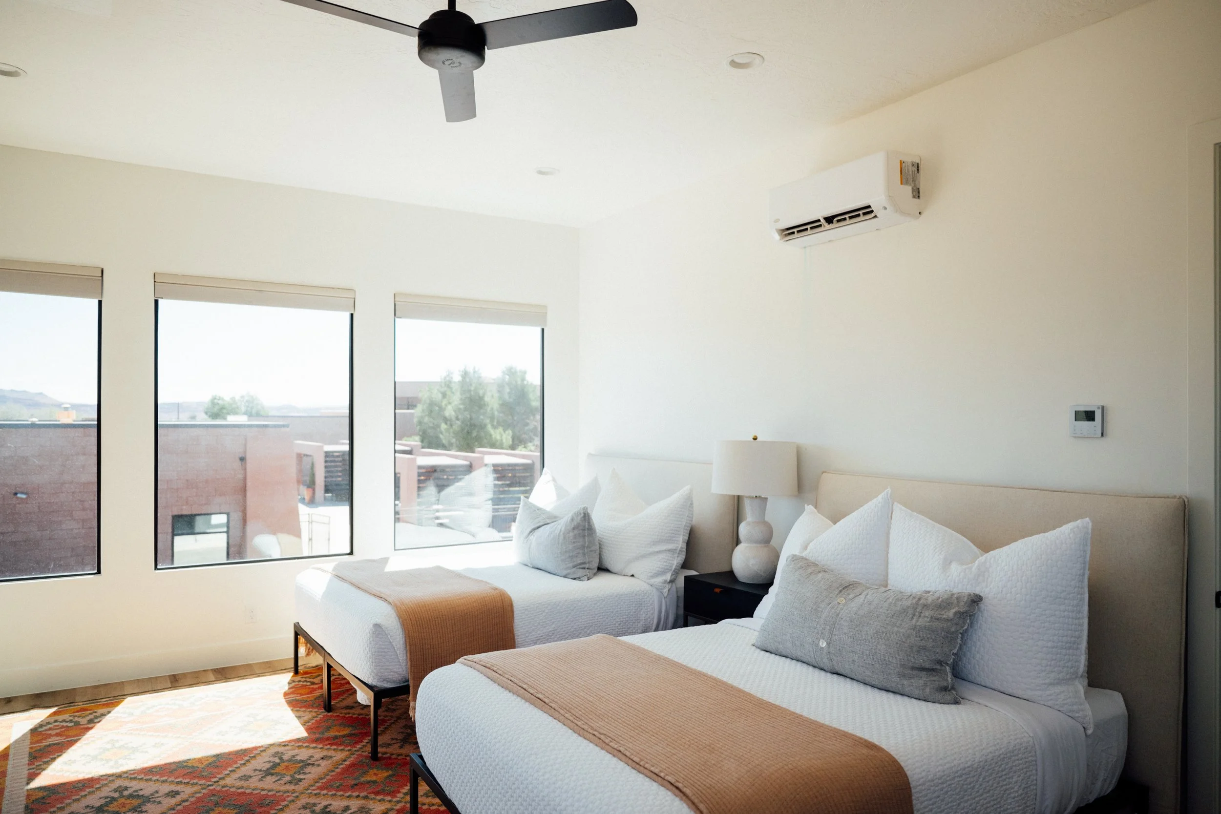 Movara Villa Double Queen Room with bright large windows, a ceiling fan, wall-mounted air conditioner, and white bedding and decorative pillows.