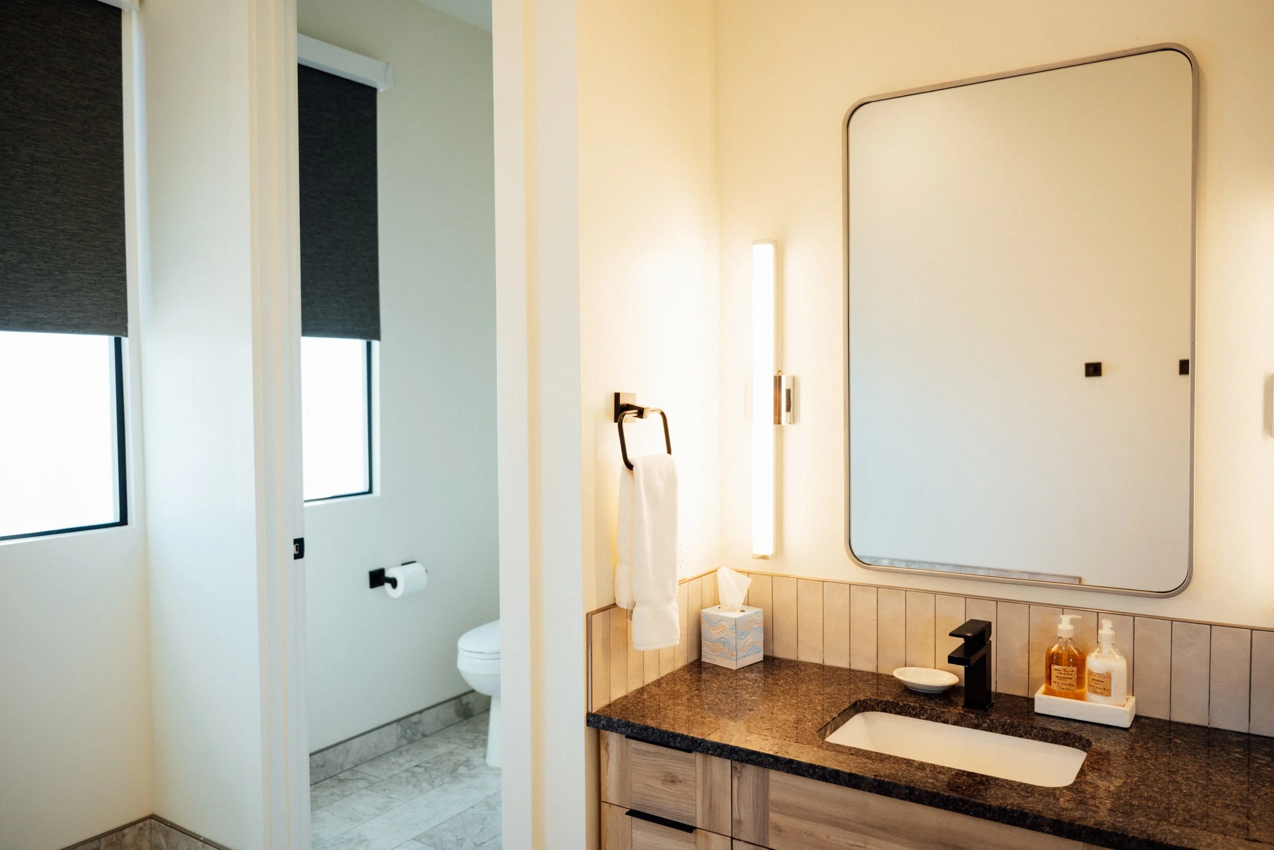 A modern bathroom with a large mirror above a granite countertop and a white sink. The countertop has soap and lotion dispensers. There is a wall-mounted towel ring with a white towel, and a toilet is visible behind partial wall separating it from th