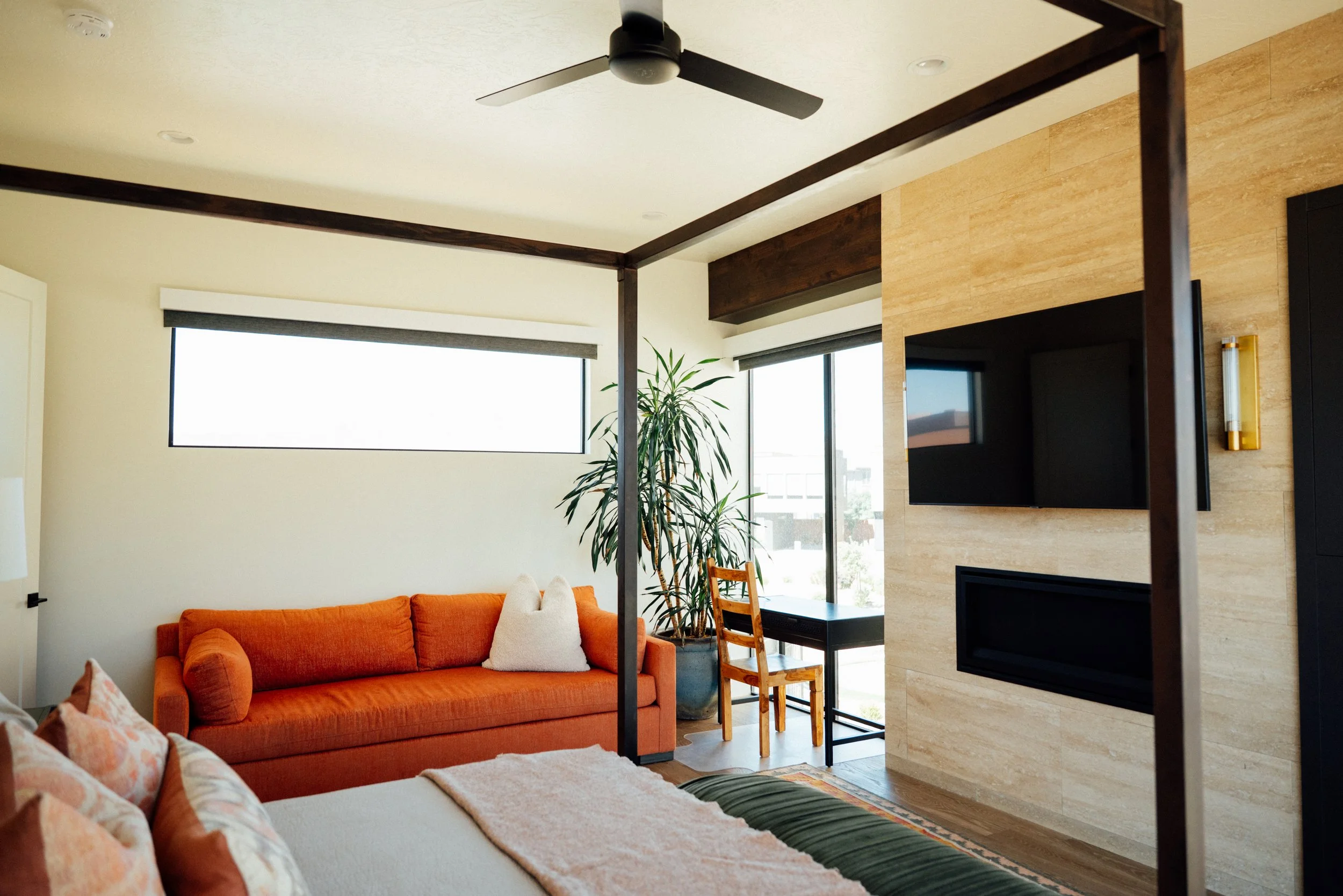 Movara villa primary suite with a modern living room with an orange sofa, cream walls, large windows with blinds, a tall plant, a black wall-mounted TV, a wooden dining chair, and a ceiling fan.