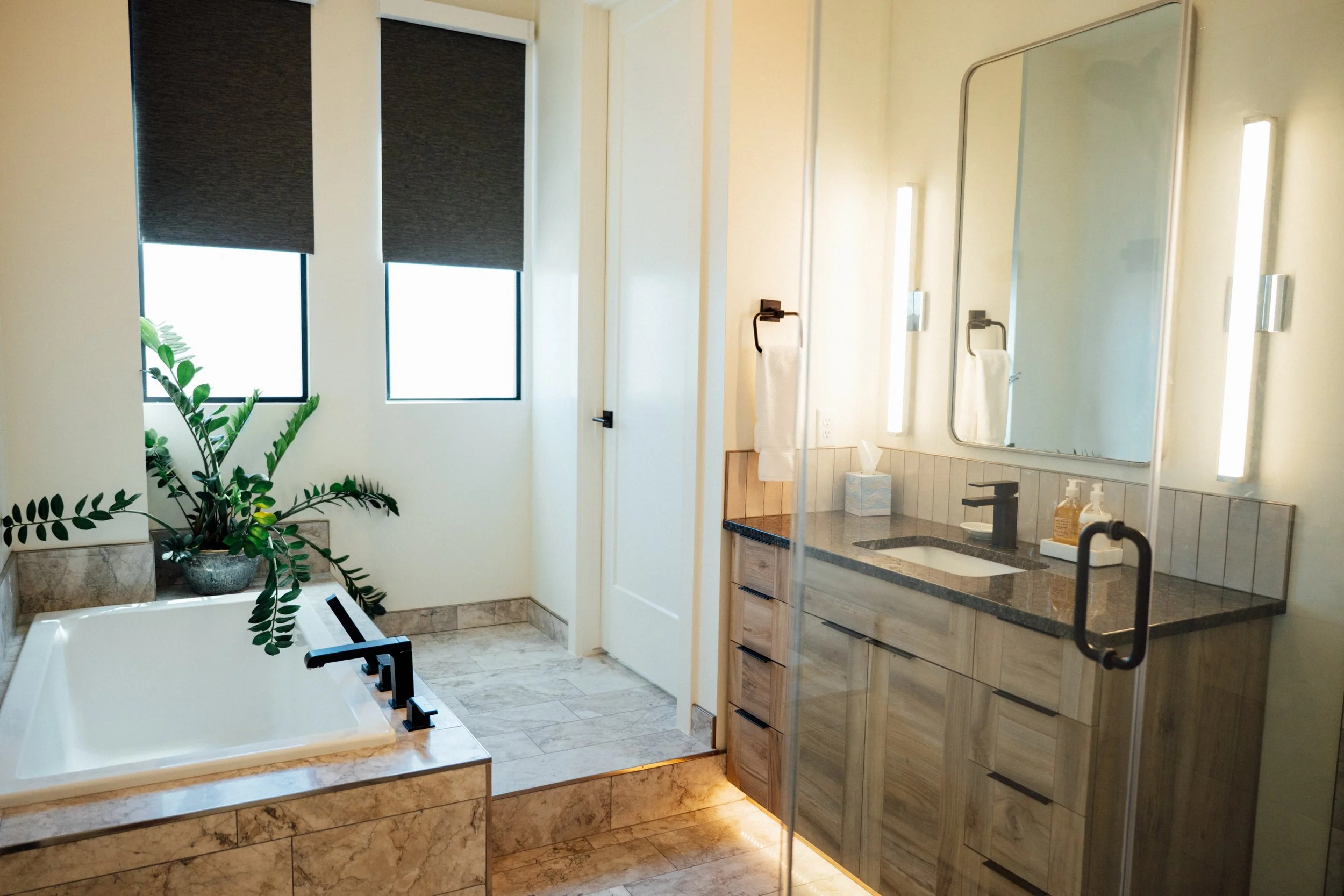 Bathroom with a bathtub, two windows with dark blinds, a large mirror, a countertop with a sink, hand soap, tissue box, and a towel on a rack. Inside the shower, a door with a handle is visible.