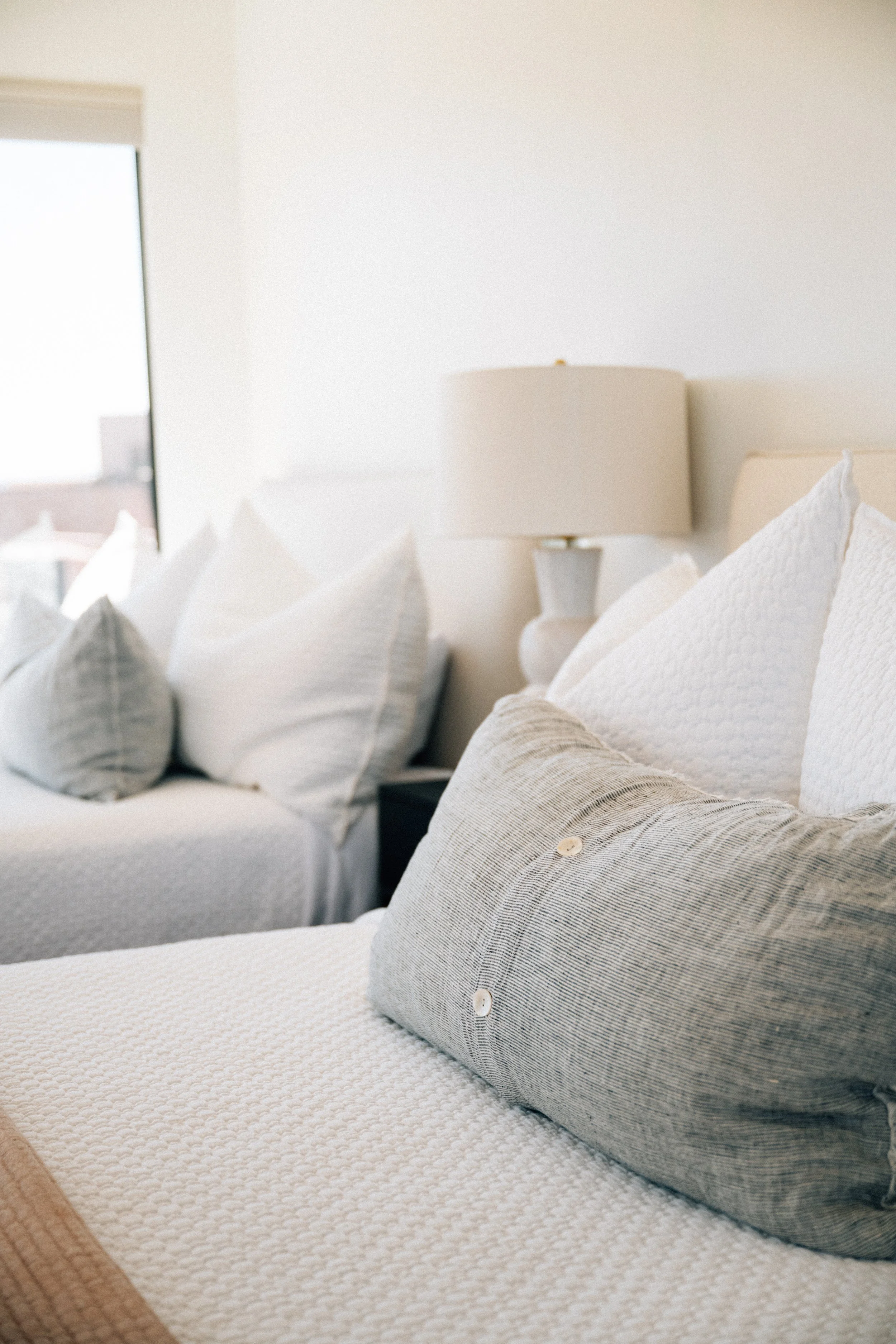 Close-up of a bed with white textured bedspread, multiple white pillows, and a striped gray and white pillow in the foreground. A beige bedside lamp is visible on the nightstand, and a window with partially open blinds is in the background, letting i