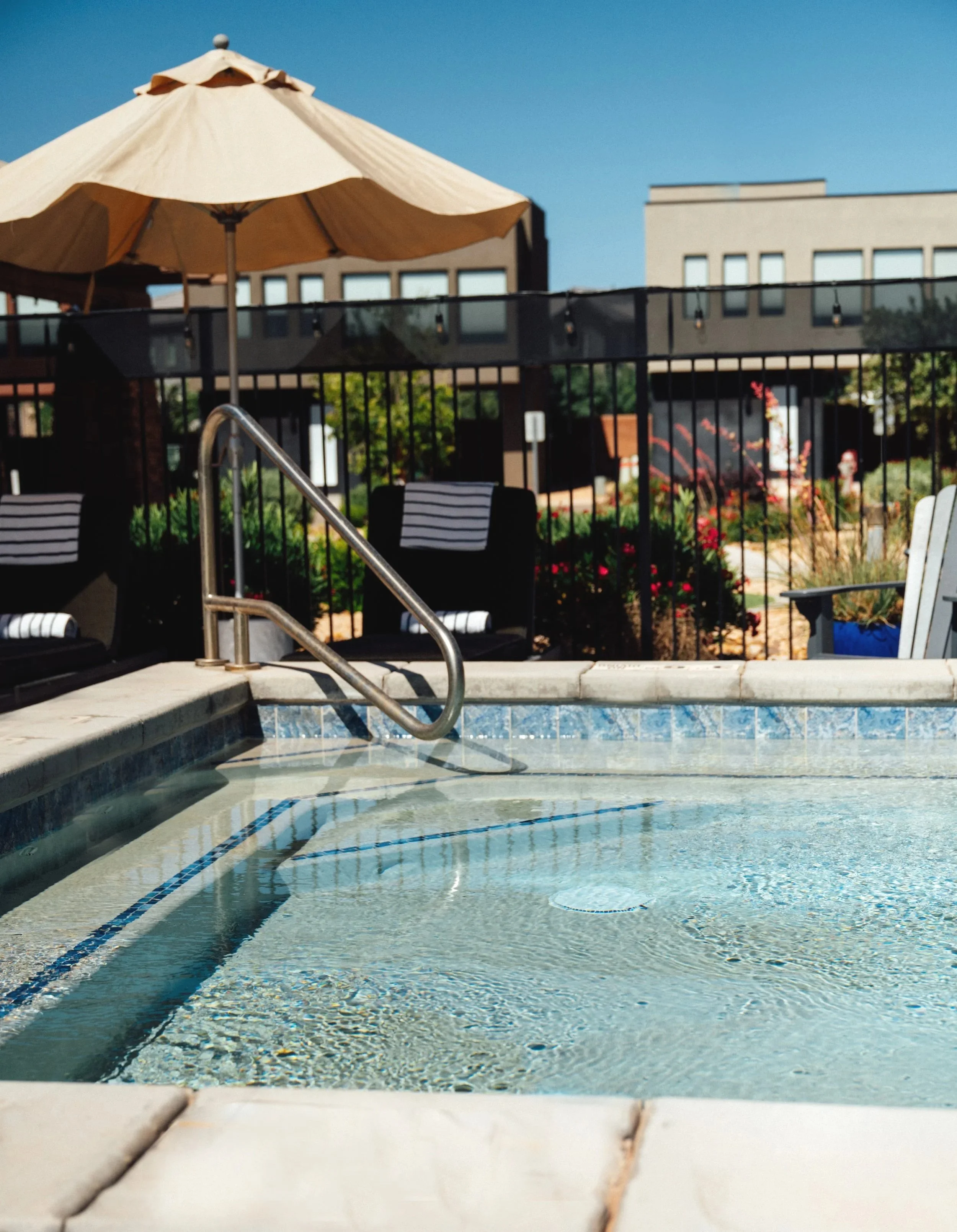 Outdoor dining area with a set table under a canopy, featuring a ceiling fan and string lights. Large windows offer a view of a pool and buildings.
