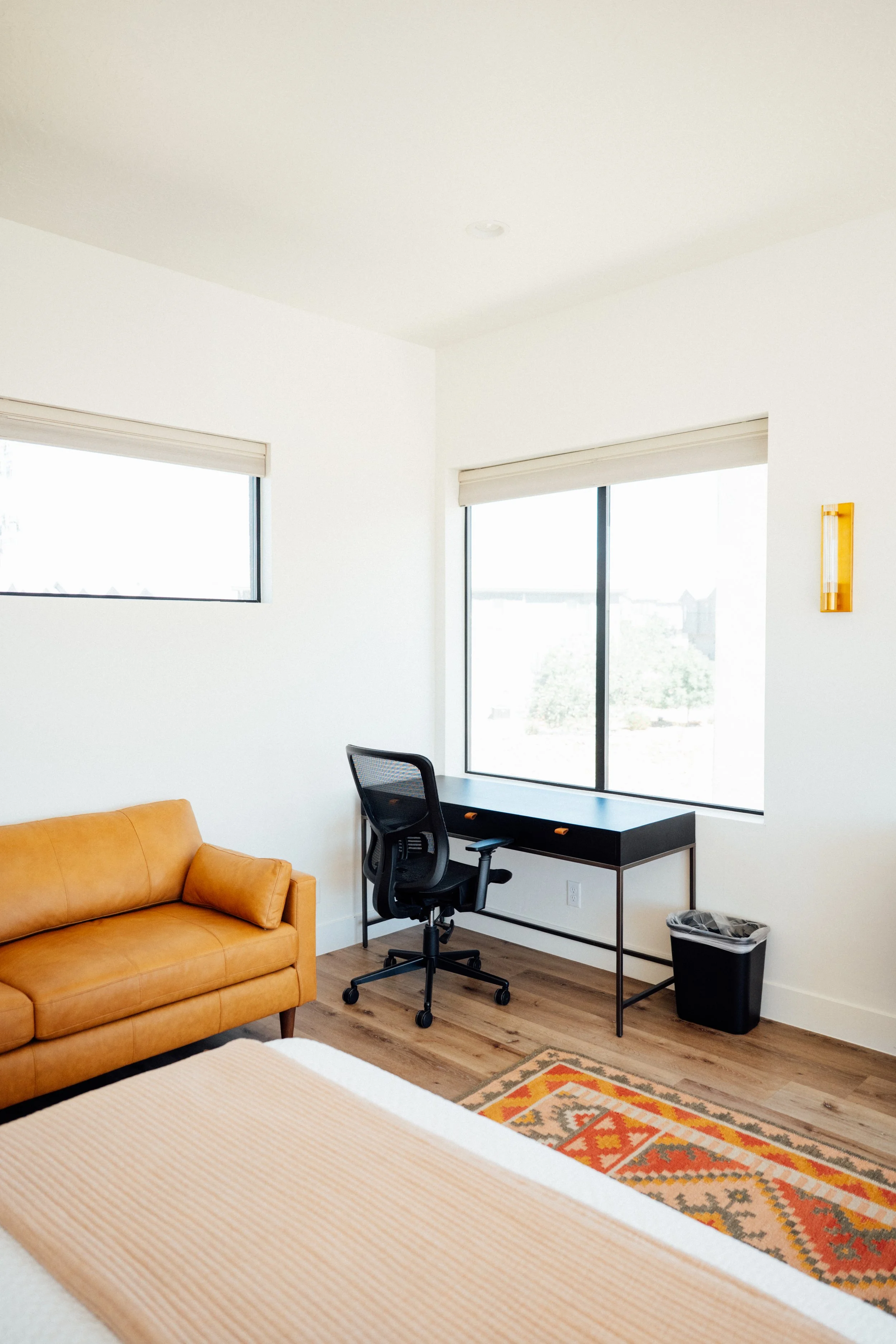 A bedroom with a tan leather sofa in the foreground, a black desk and ergonomic chair near large windows with white blinds, a colorful rug on wooden floor, and a wall-mounted light fixture.