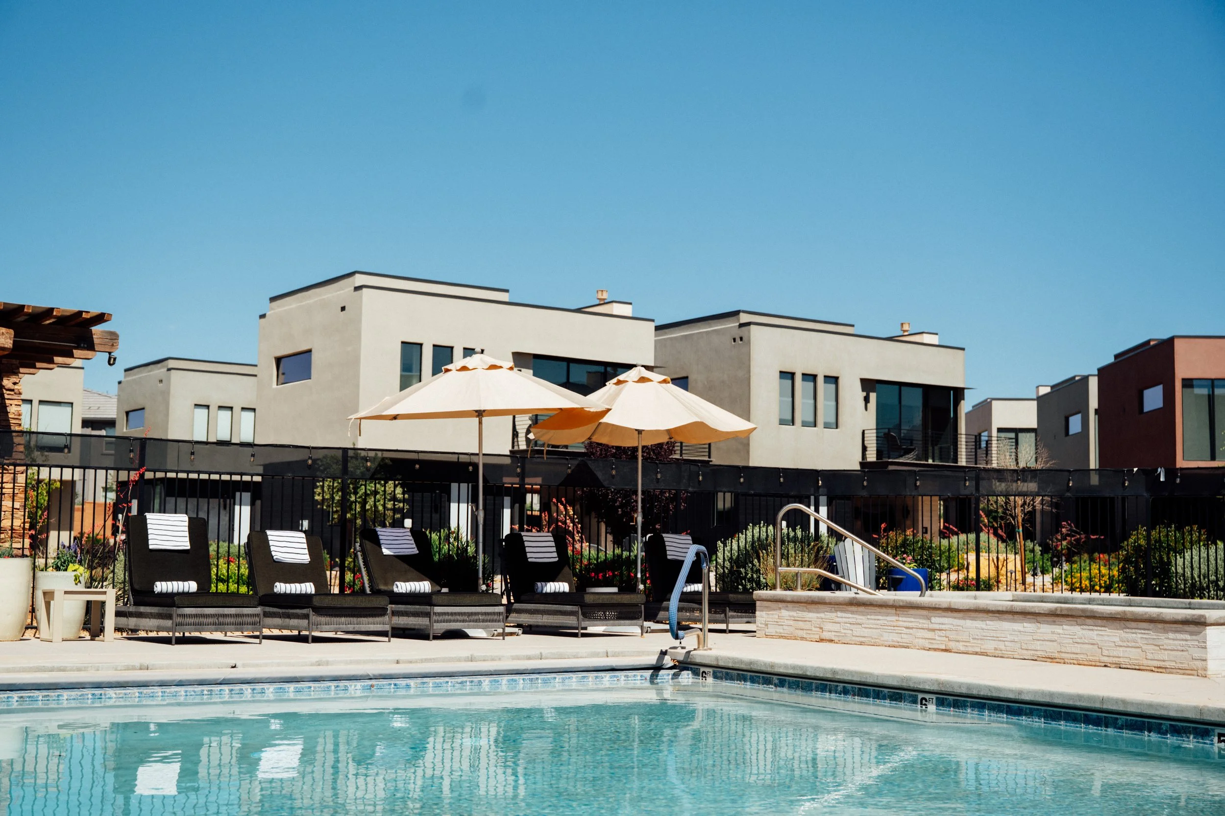 View of a backyard pool area with lounge chairs, umbrellas, and a garden, with modern multi-story apartment buildings in the background under a clear blue sky.