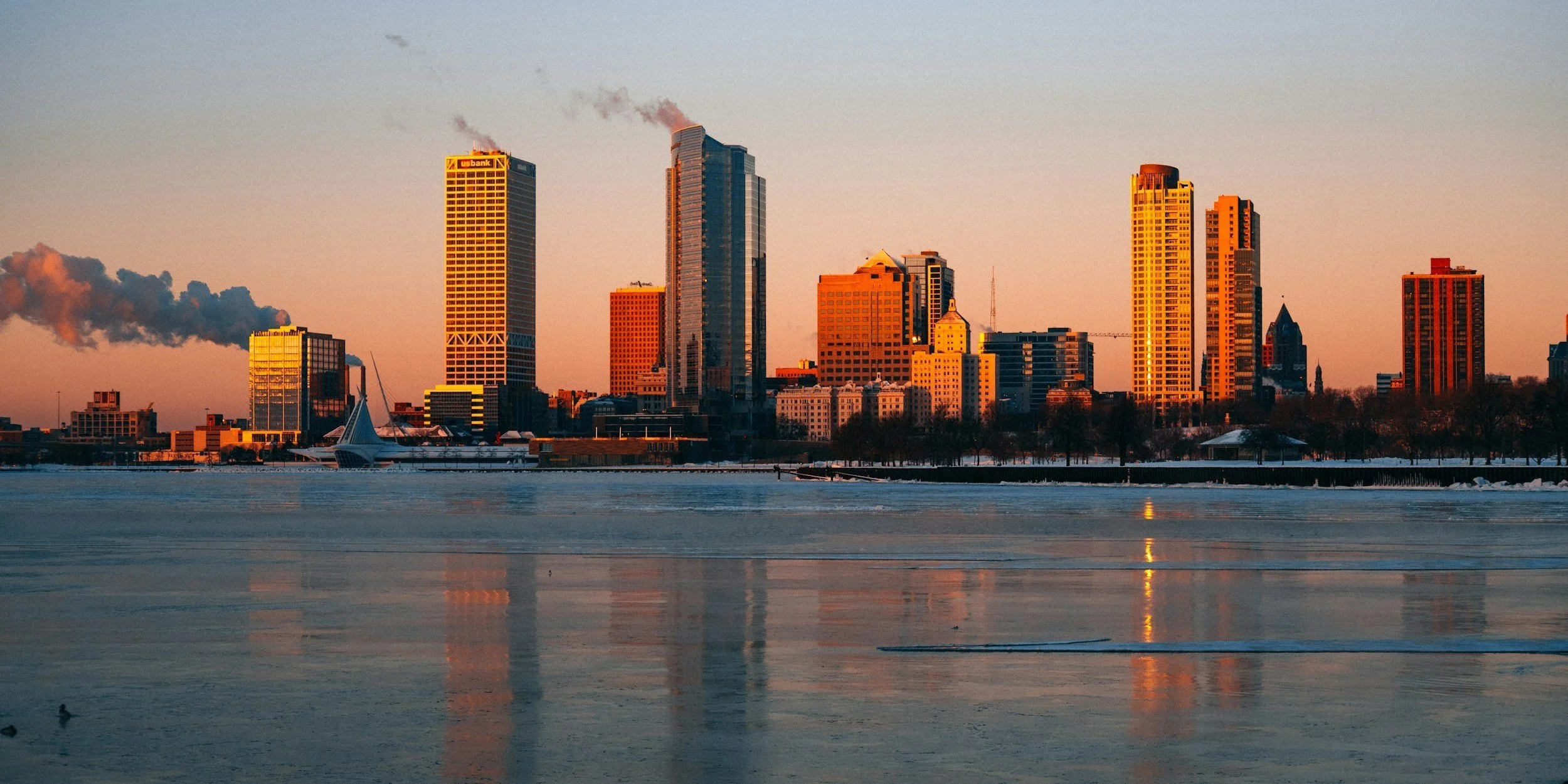 City skyline with tall buildings reflecting on a frozen body of water during sunset, with some smoke or steam rising from chimneys.