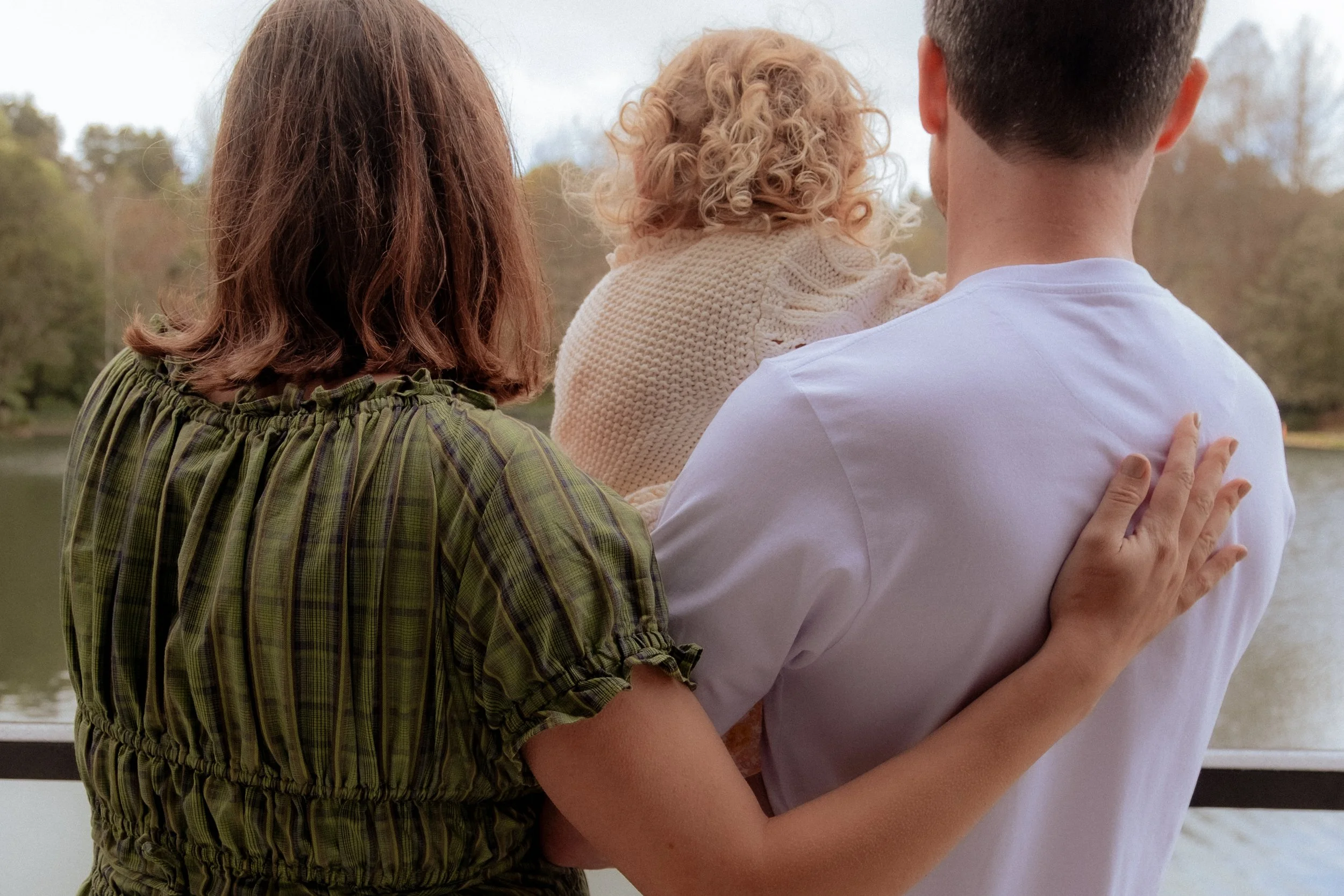 A family of three, a woman, a man, and a young girl, standing outdoors near a lake, with trees in the background. The woman has shoulder-length brown hair, the man has short dark hair, and the girl has curly blonde hair. The woman has her arm around 